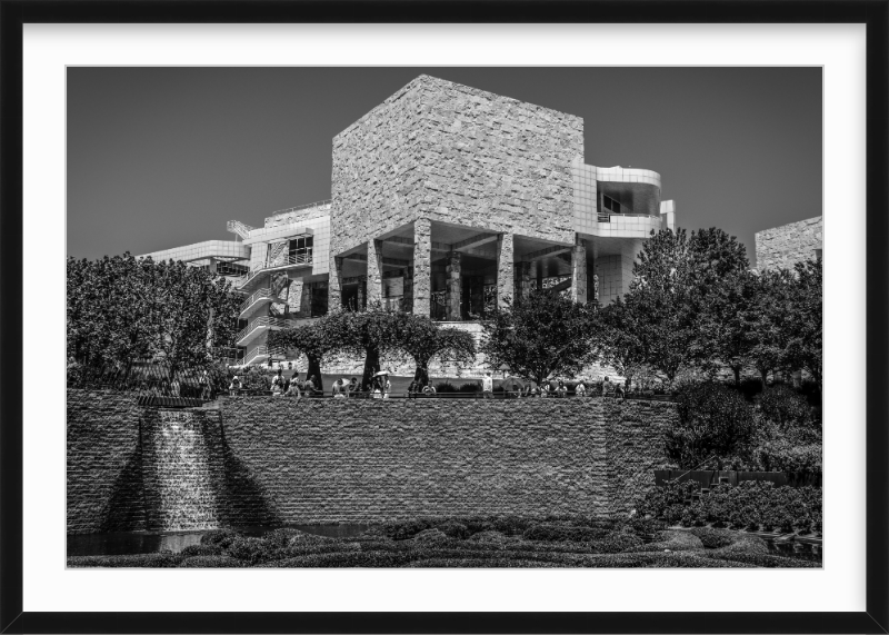 Black and white photo of a modern building with large stone blocks, supported by pillars, with trees in front and a brick wall in the foreground.