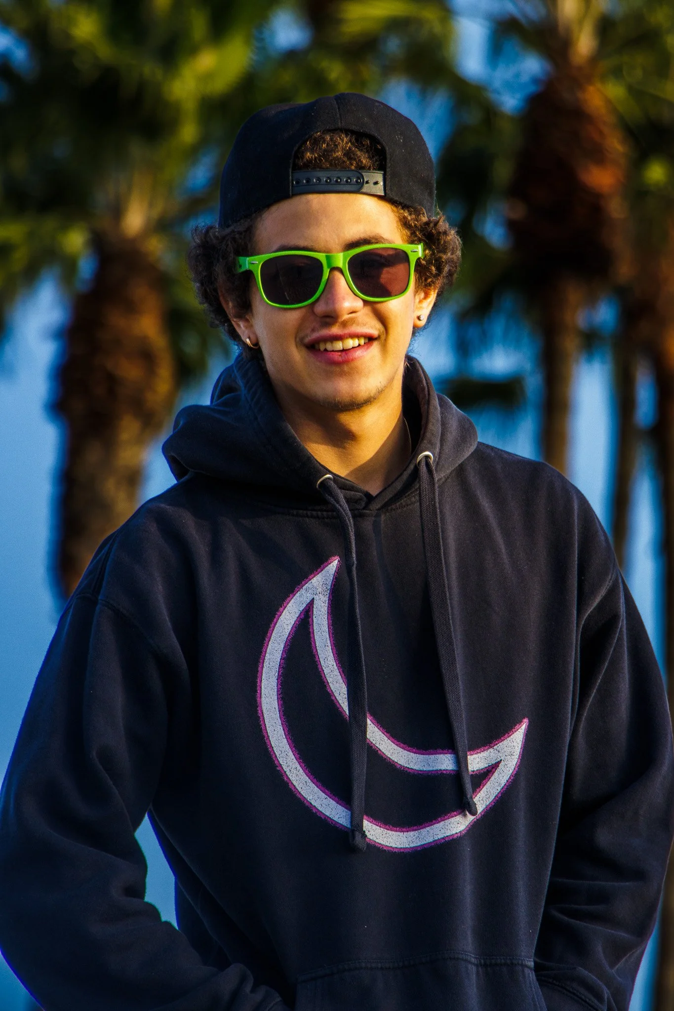 Young man wearing a black hoodie with a pink and white logo, bright green sunglasses, a black cap worn backwards, and small earrings, standing outdoors near palm trees during sunset.
