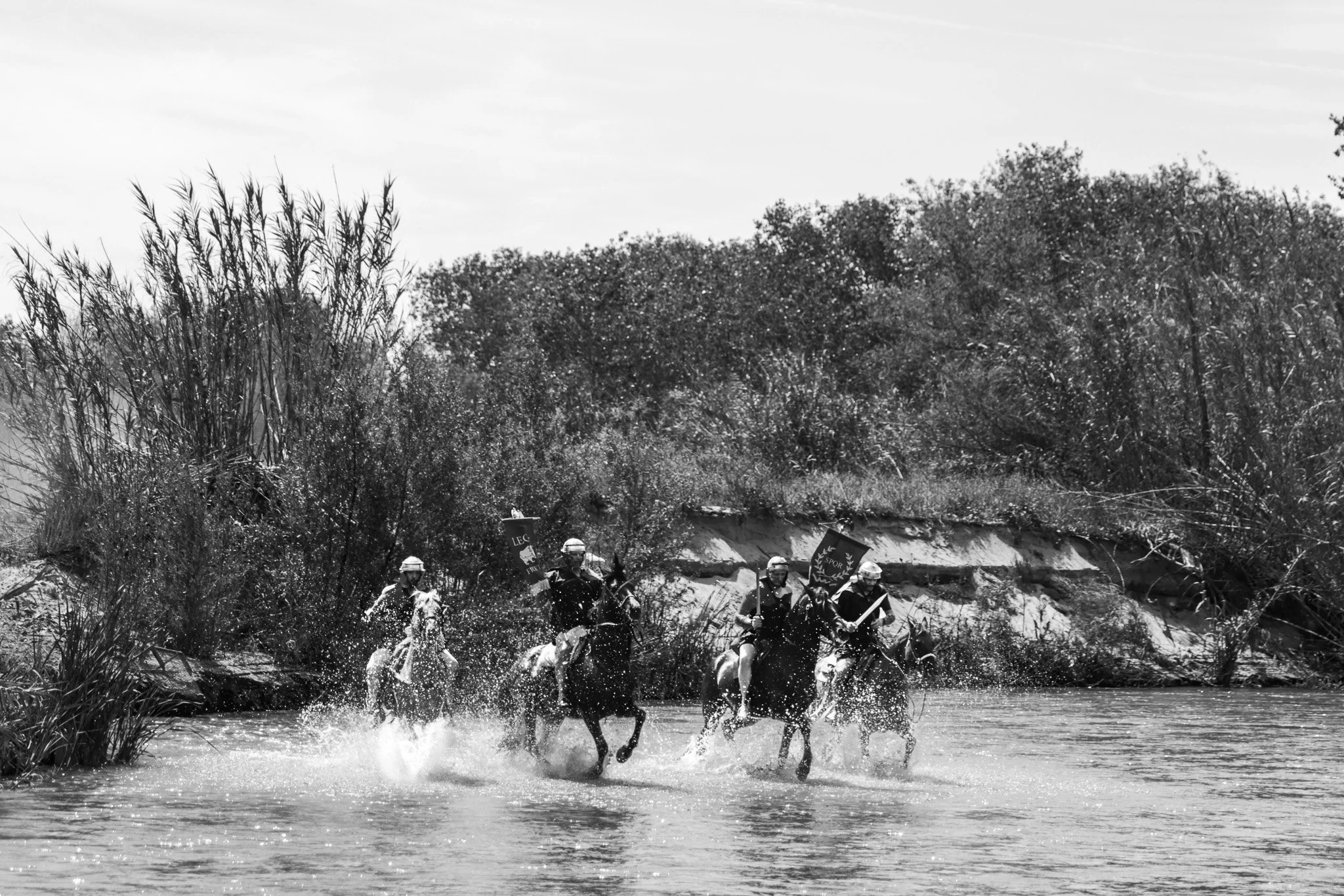 A group of soldiers on horseback crossing a river with trees and bushes in the background.