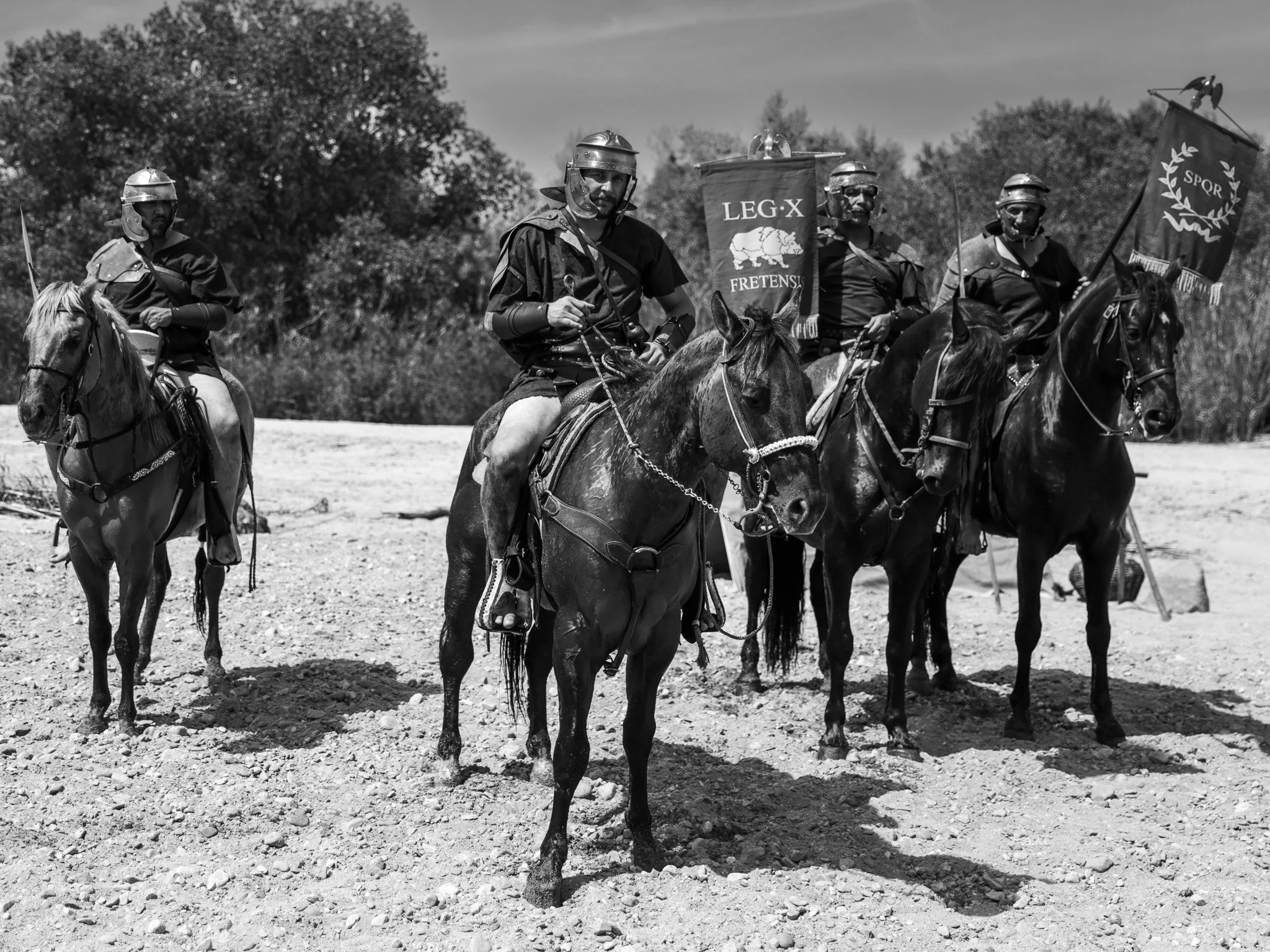 A group of men dressed as Roman soldiers on horseback, holding banners, riding on a dirt field with trees in the background.