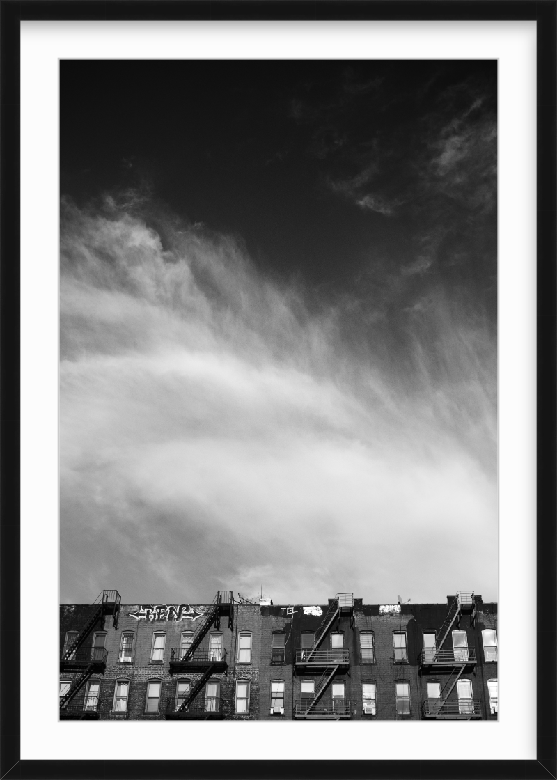 Black and white photo of an apartment building with fire escapes and graffiti on the rooftop, under a cloudy sky.
