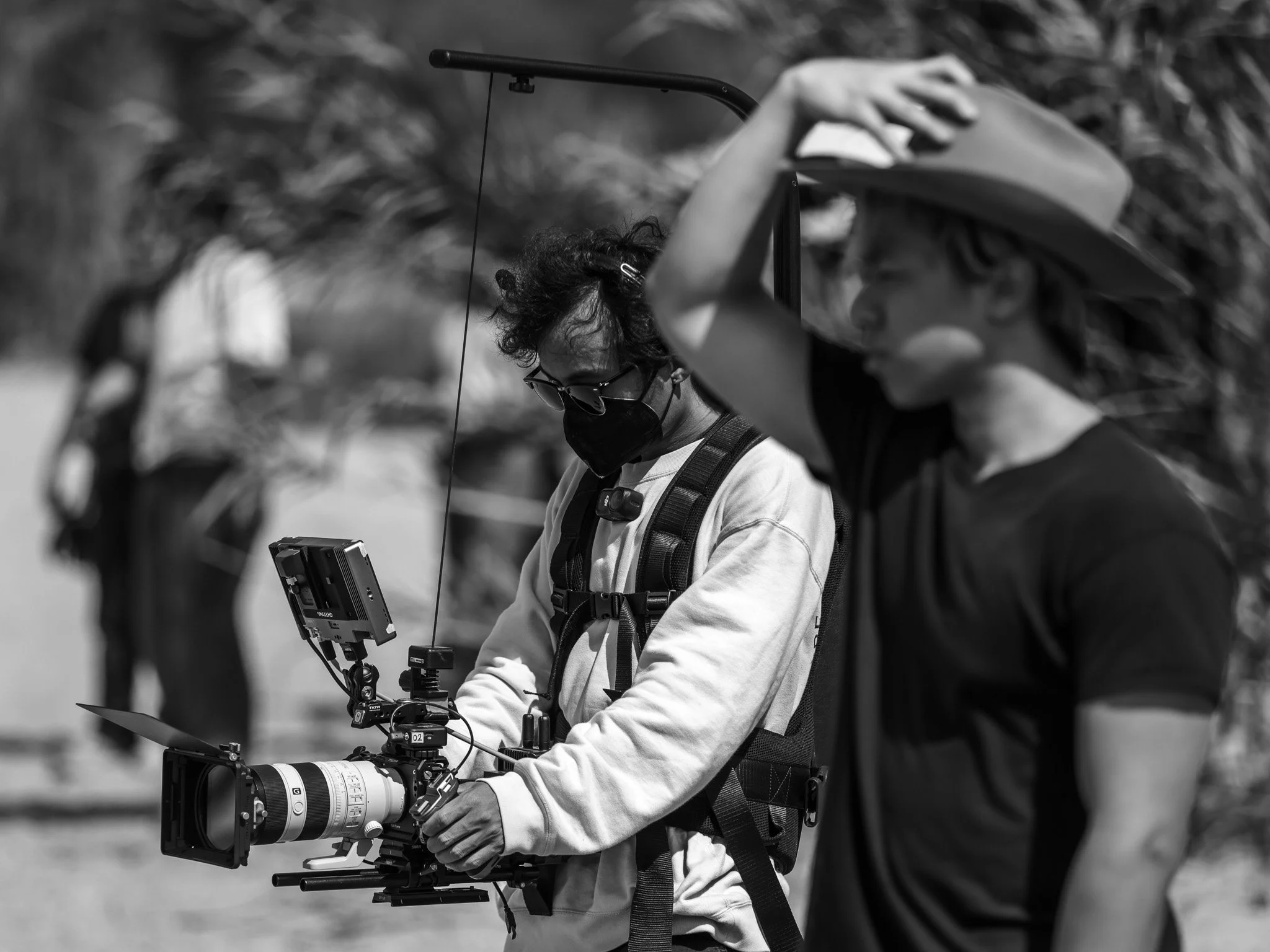 A person wearing glasses and a face mask operating a large camera with a telephoto lens, outdoors with blurred foliage in the background.