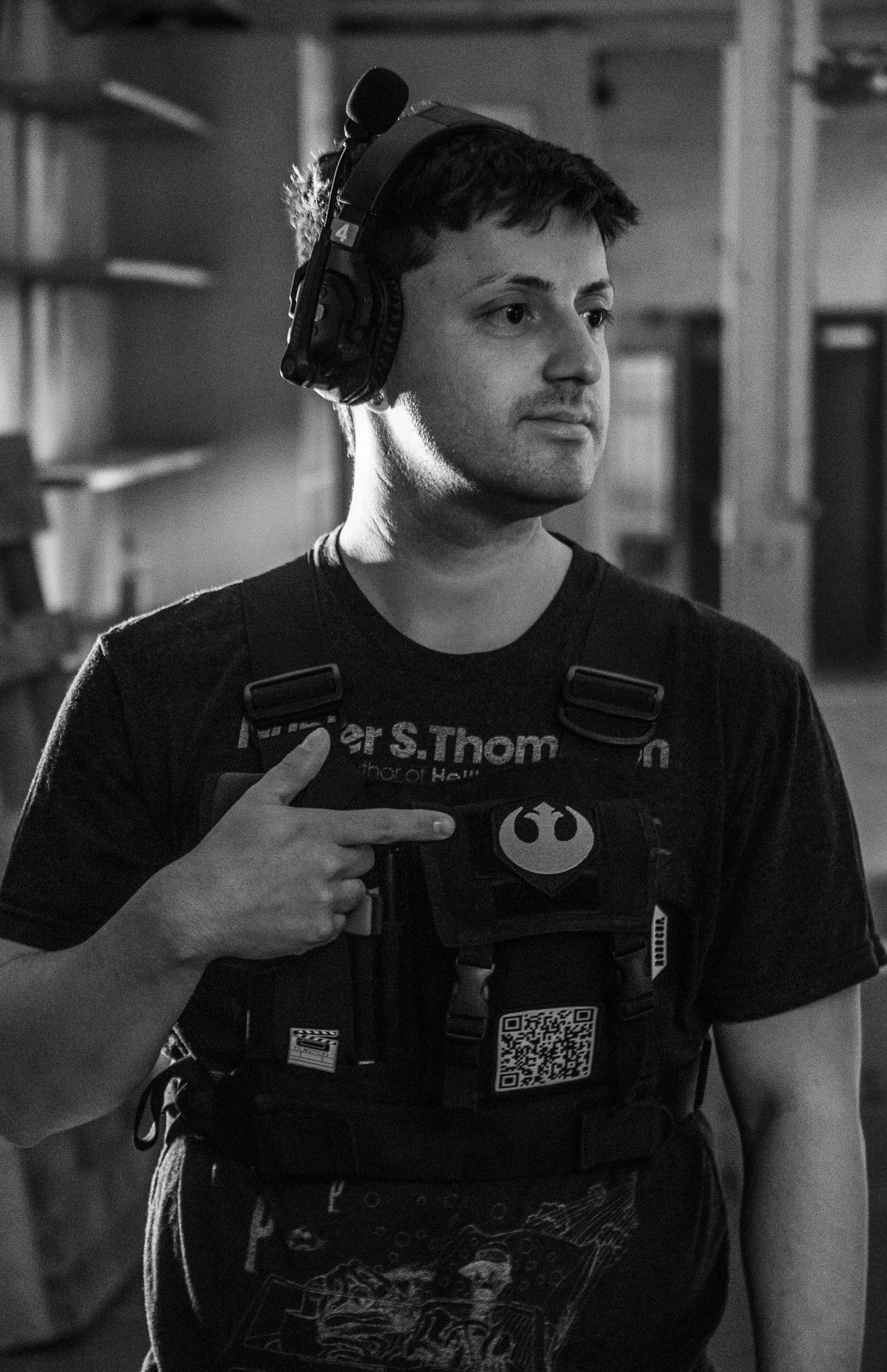 A young man taking a mirror selfie in black and white, wearing a Star Wars-themed T-shirt, a vest with a QR code, and pointing to the insignia on his vest.