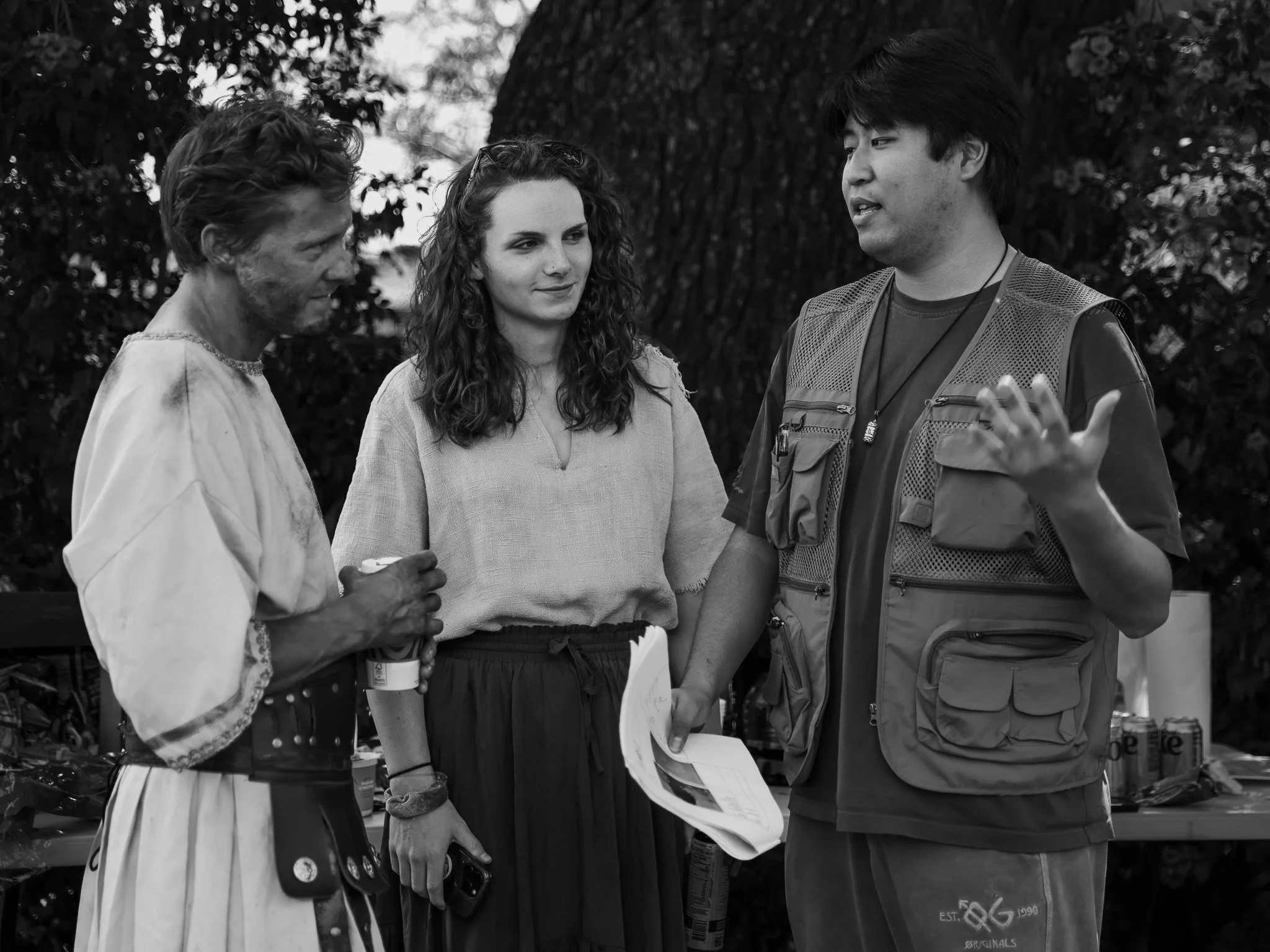 Three people in conversation outdoors, two men and a woman, with trees and a food stand in the background, black and white photograph.