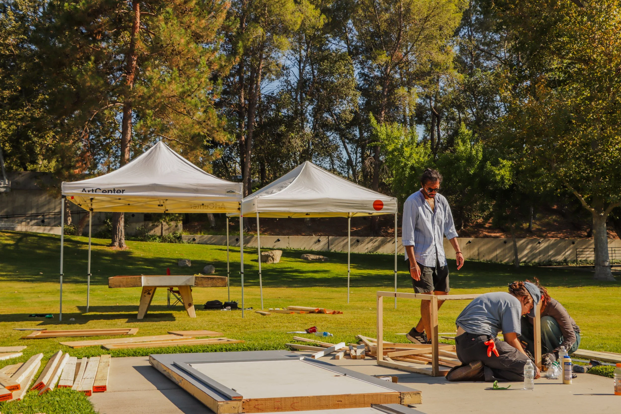 People assembling wooden structures outdoors under trees with white tents in the background.