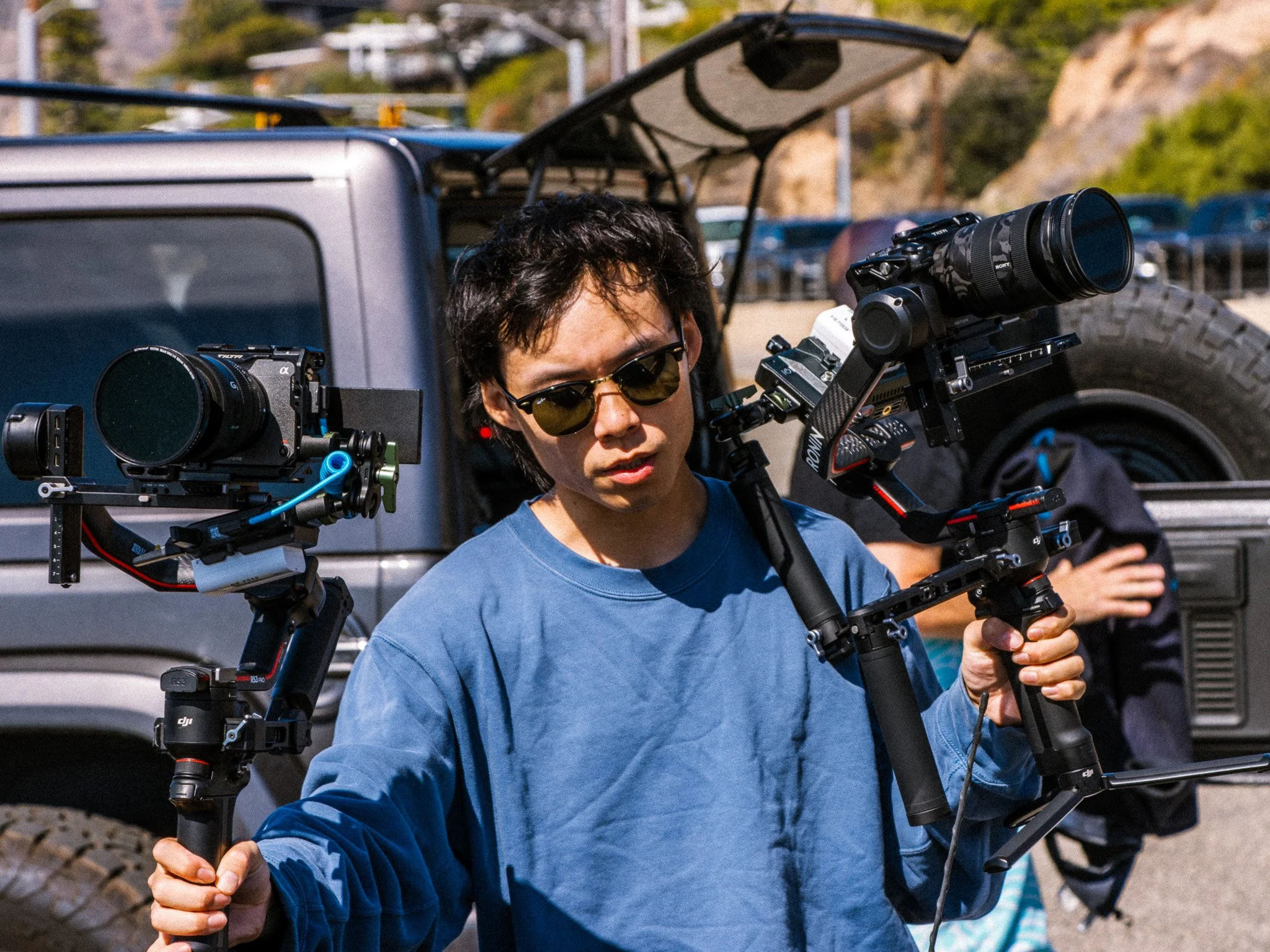 A person with short, dark hair wearing sunglasses and a blue long-sleeve shirt operating a professional camera mounted on a stabilizer rig outdoors. Another person is visible in the background near a vehicle.