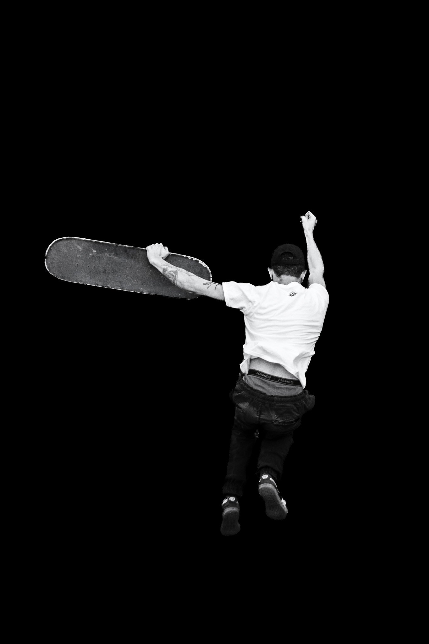 A person with tattoos on their right arm wearing a white t-shirt, black cargo pants, a backward baseball cap, and sneakers, captured mid-air holding a skateboard, against a black background.