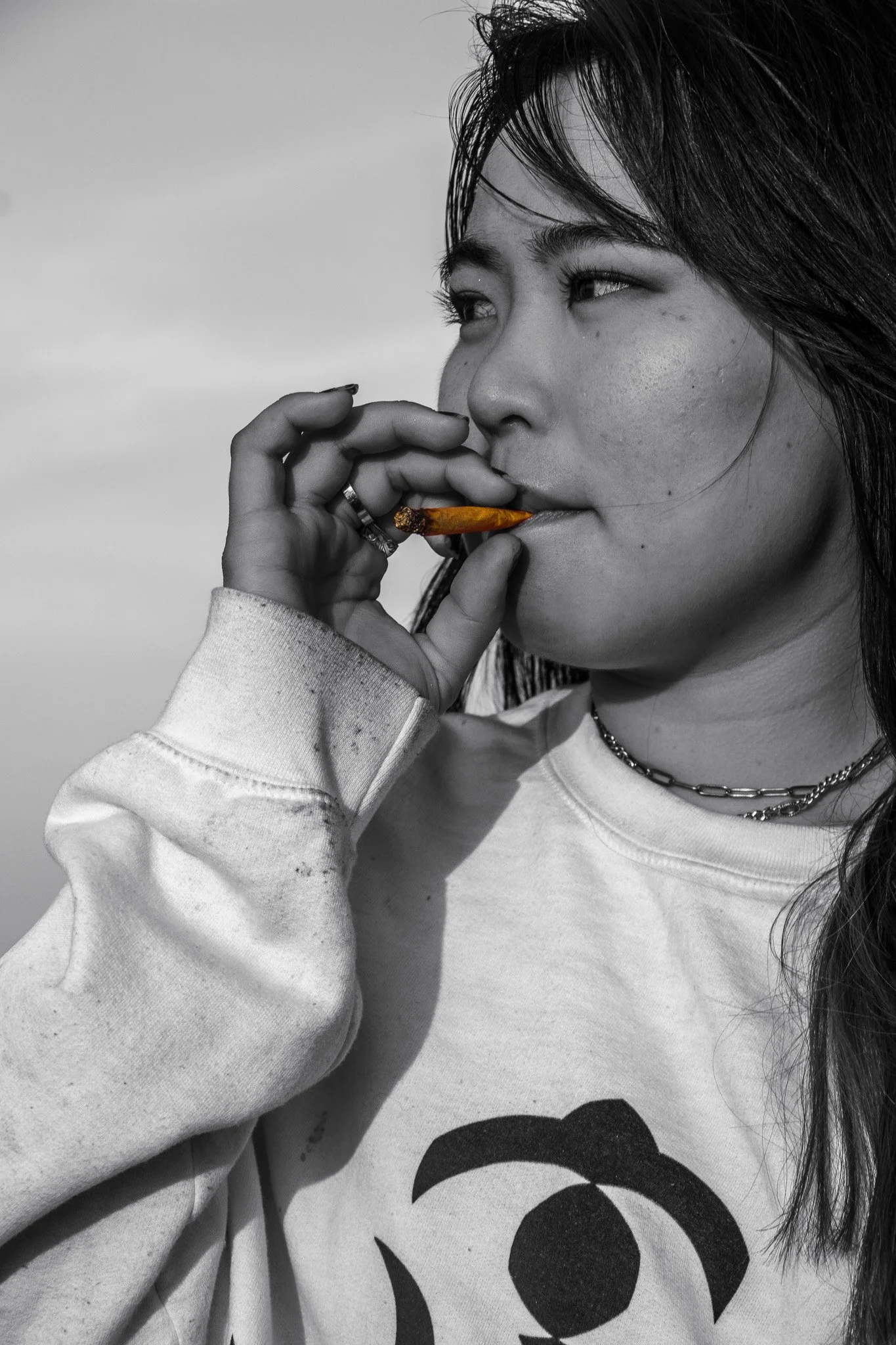 A woman with long hair holding a cigar in her fingers near her lips, wearing a white sweatshirt with black abstract design, and layered chains around her neck, against a cloudy sky background.