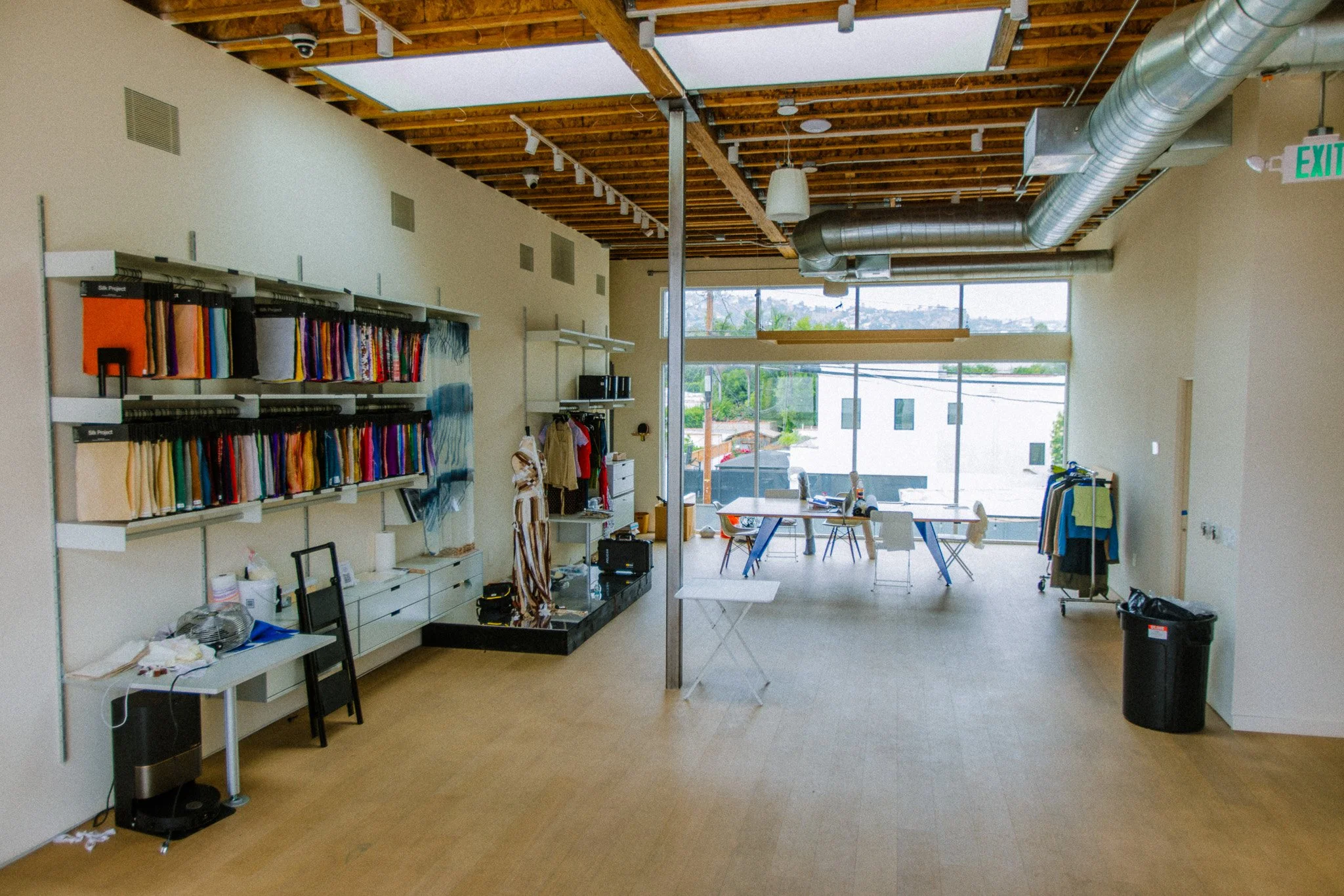 Interior view of a modern commercial or office space with furniture, fabric samples on shelves, a table, chairs, large windows, and an exposed wooden ceiling with ventilation ducts.