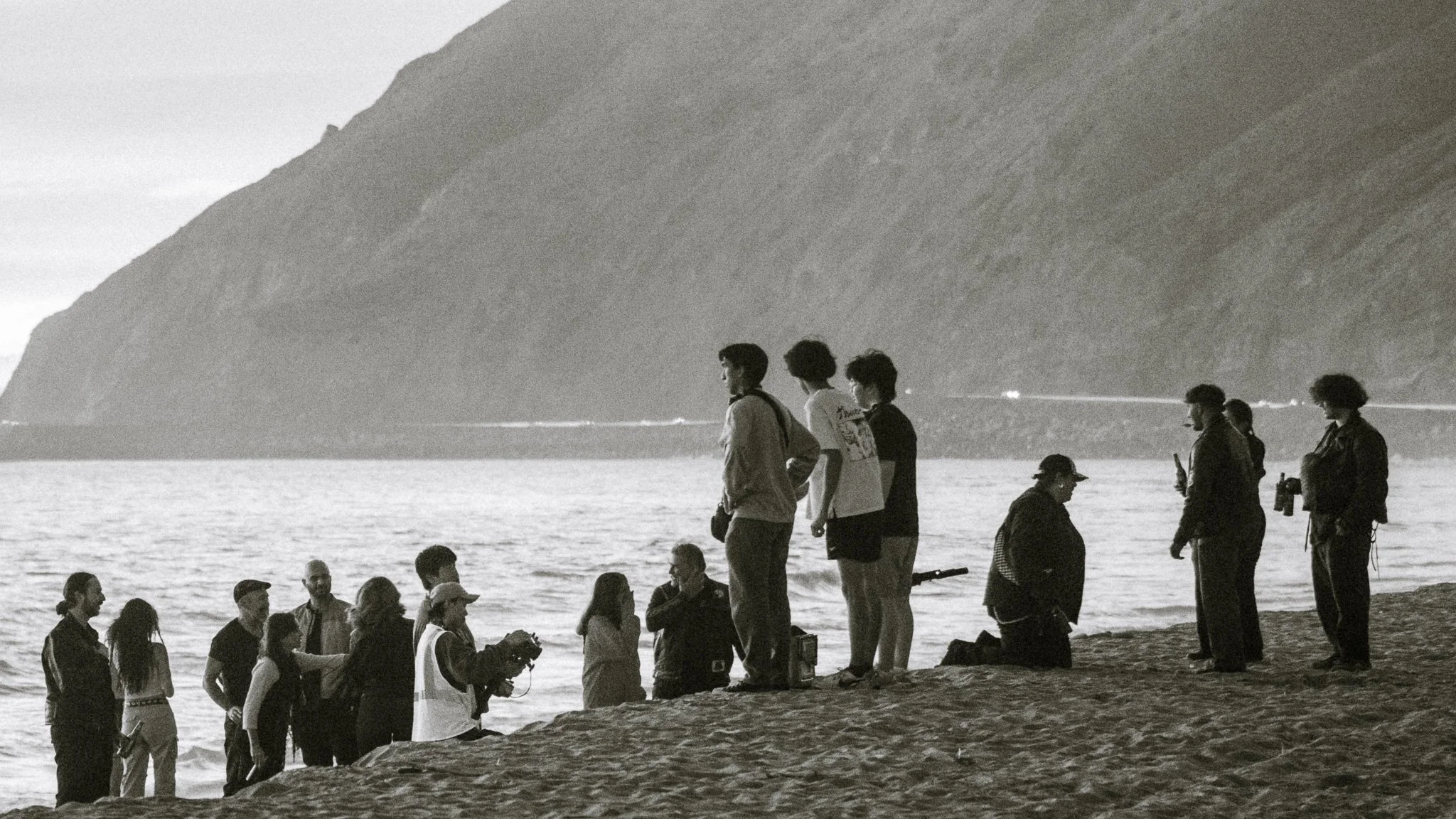 Group of people standing and sitting on a beach near the water with a mountain in the background, some looking at their phones, in black and white.