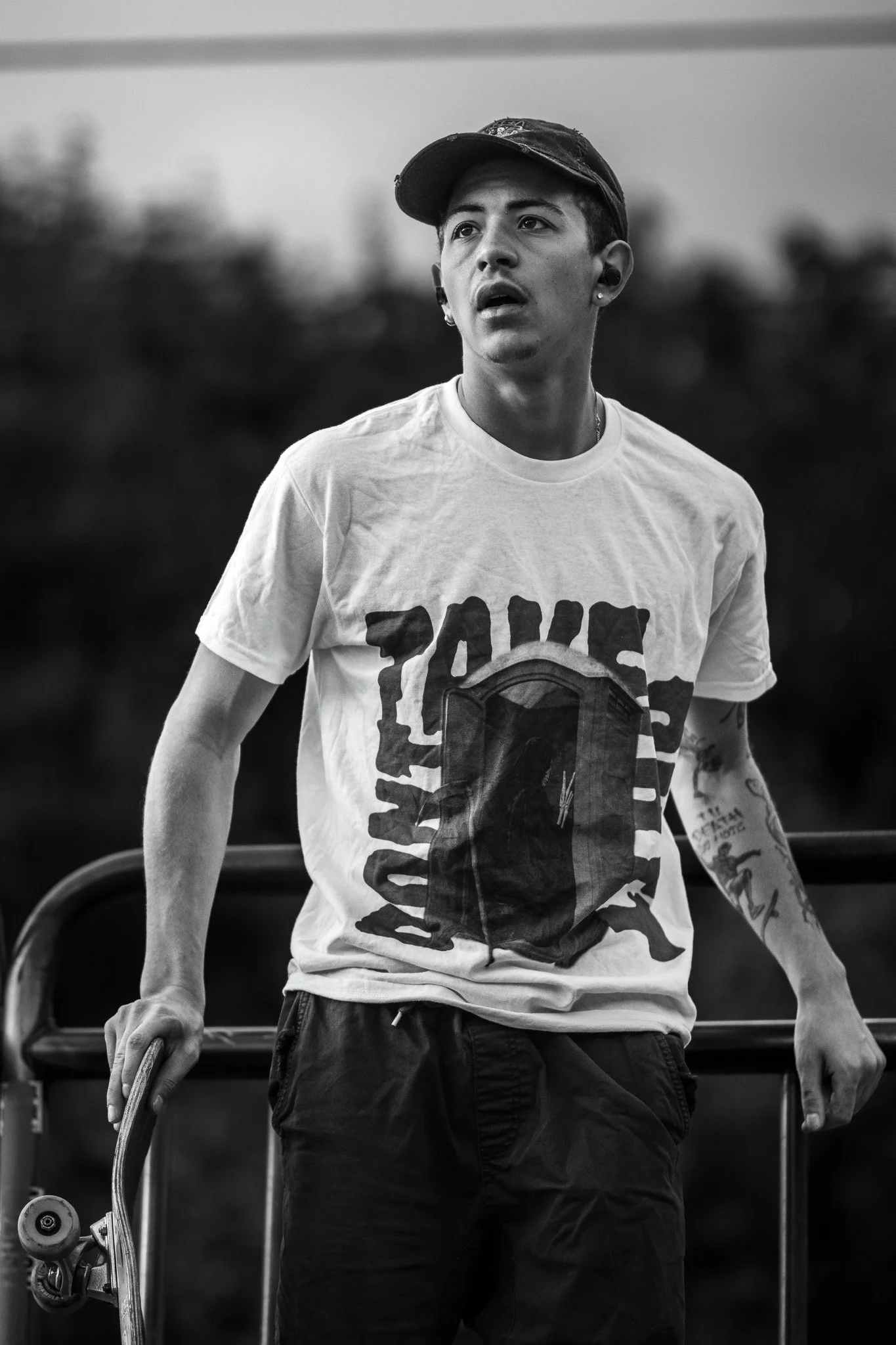 A young man in a baseball cap and t-shirt with a graphic print, holding a skateboard in his right hand, standing outdoors with a blurred background.