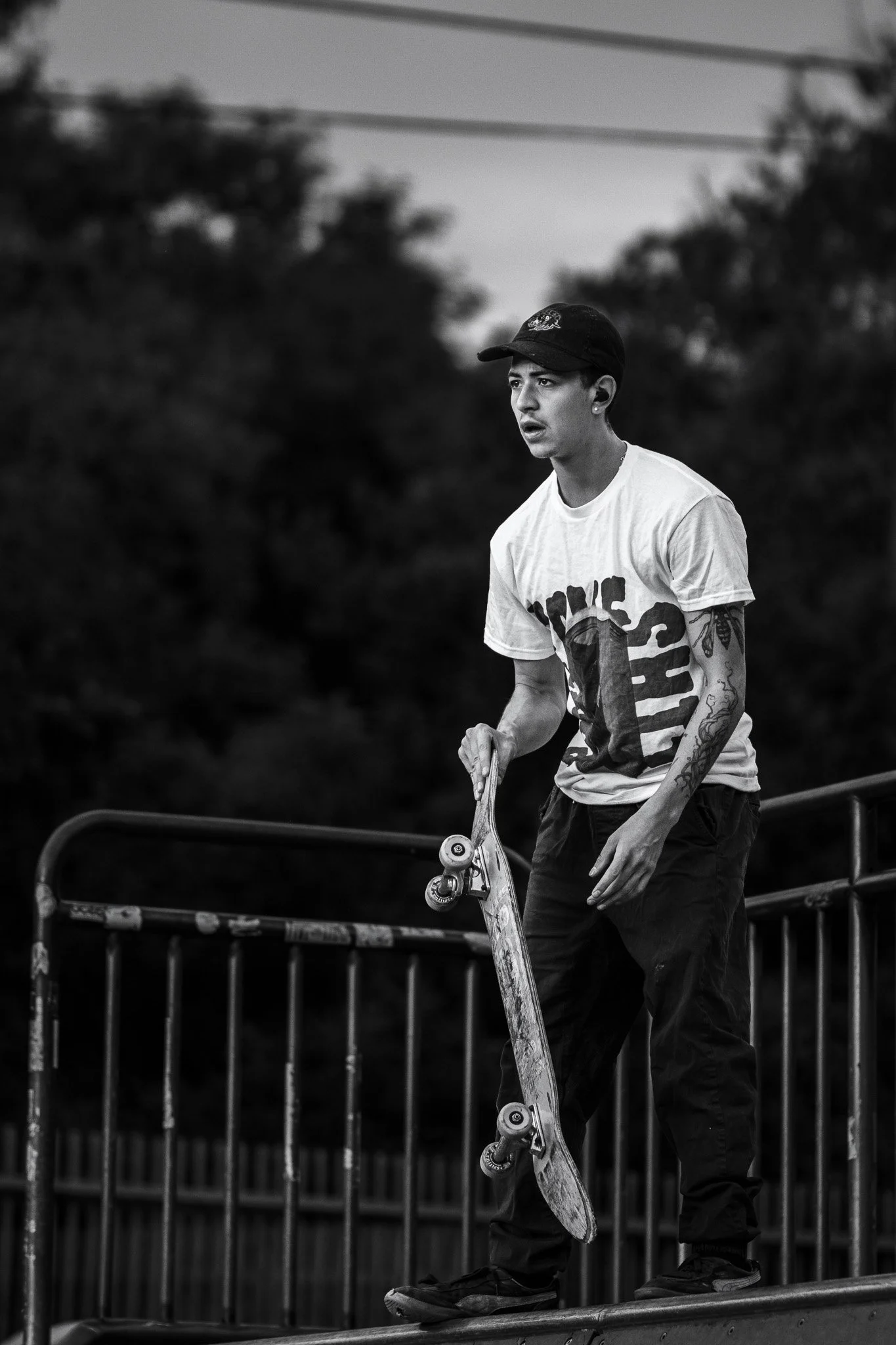 A young male skateboarder wearing a cap, T-shirt, and pants, standing on a skateboard on a ledge in an outdoor skatepark, with trees and a metal fence in the background.