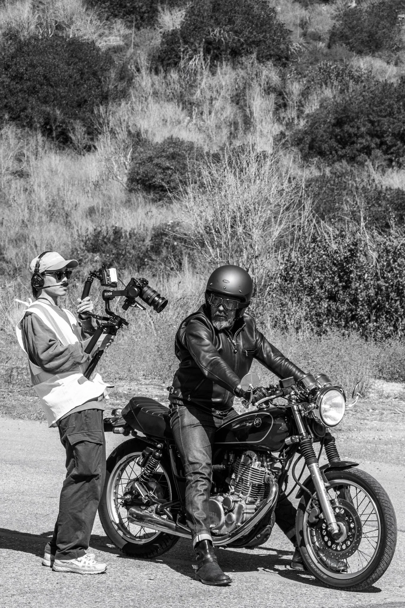 A person riding a motorcycle wearing a leather jacket and helmet, with a person standing beside them operating a camera on a gimbal, in an outdoor setting with shrubs and trees.