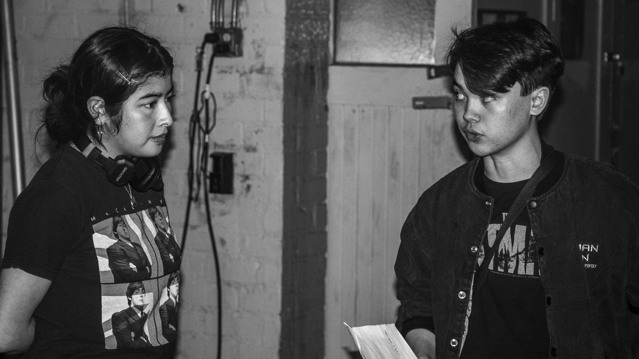 Two young people, a woman and a man, having a conversation in an industrial or warehouse-like setting. The woman is wearing a graphic t-shirt with images of a band and has headphones around her neck, while the man is dressed in a jacket with a graphi