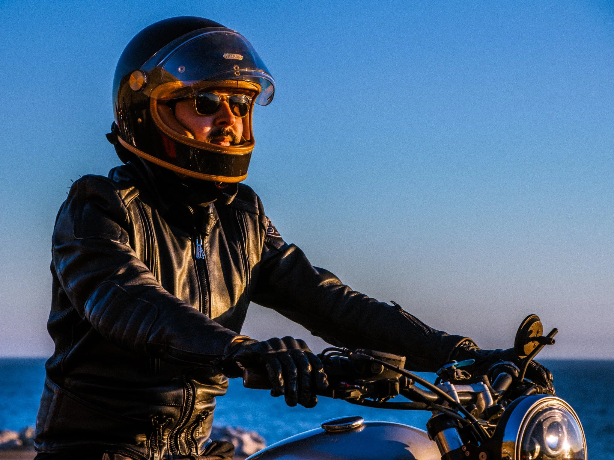 Man riding a motorcycle outdoors at sunset, wearing a black leather jacket, helmet with sunglasses, and gloves.