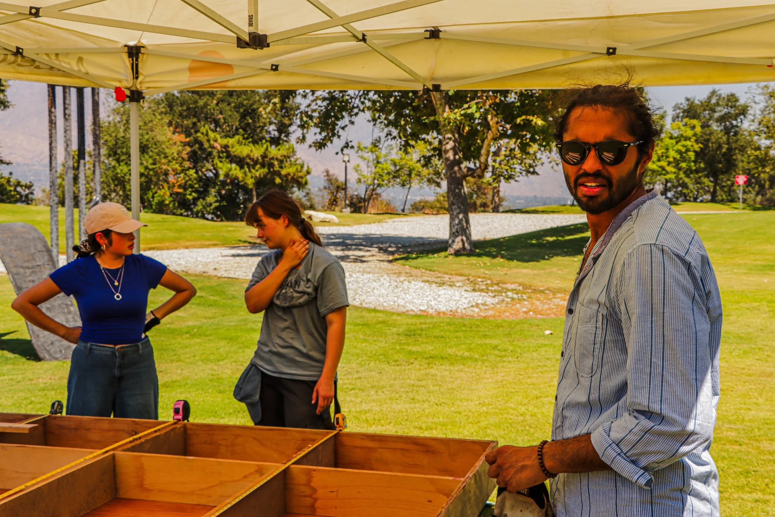 Three people outdoors under a shade tent, with a man in sunglasses looking at a wooden game or craft, two young women nearby, in a park with trees and a grassy area.
