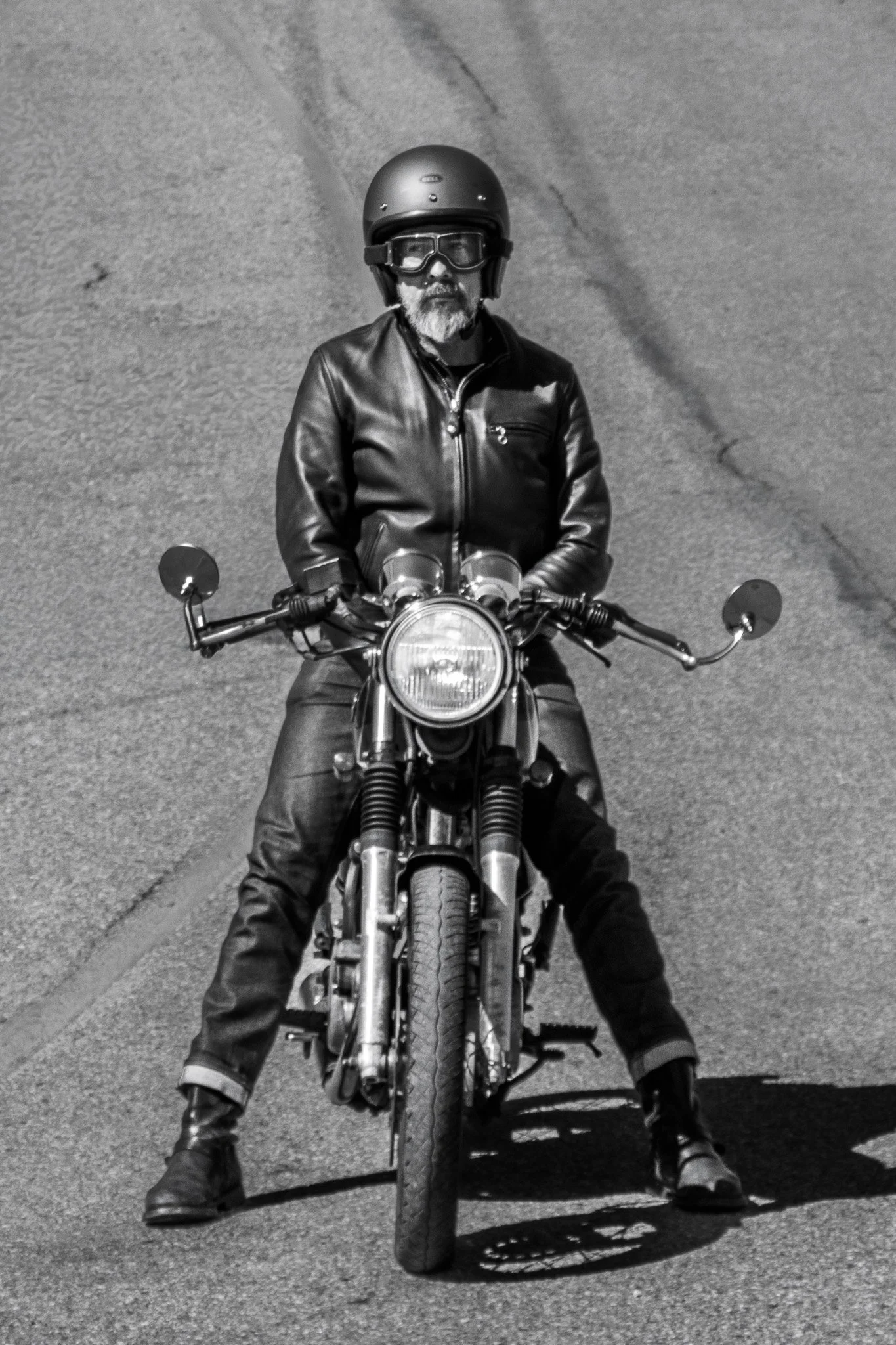 Black and white photo of a man wearing a helmet, goggles, leather jacket, and boots, sitting on a motorcycle on a paved street.