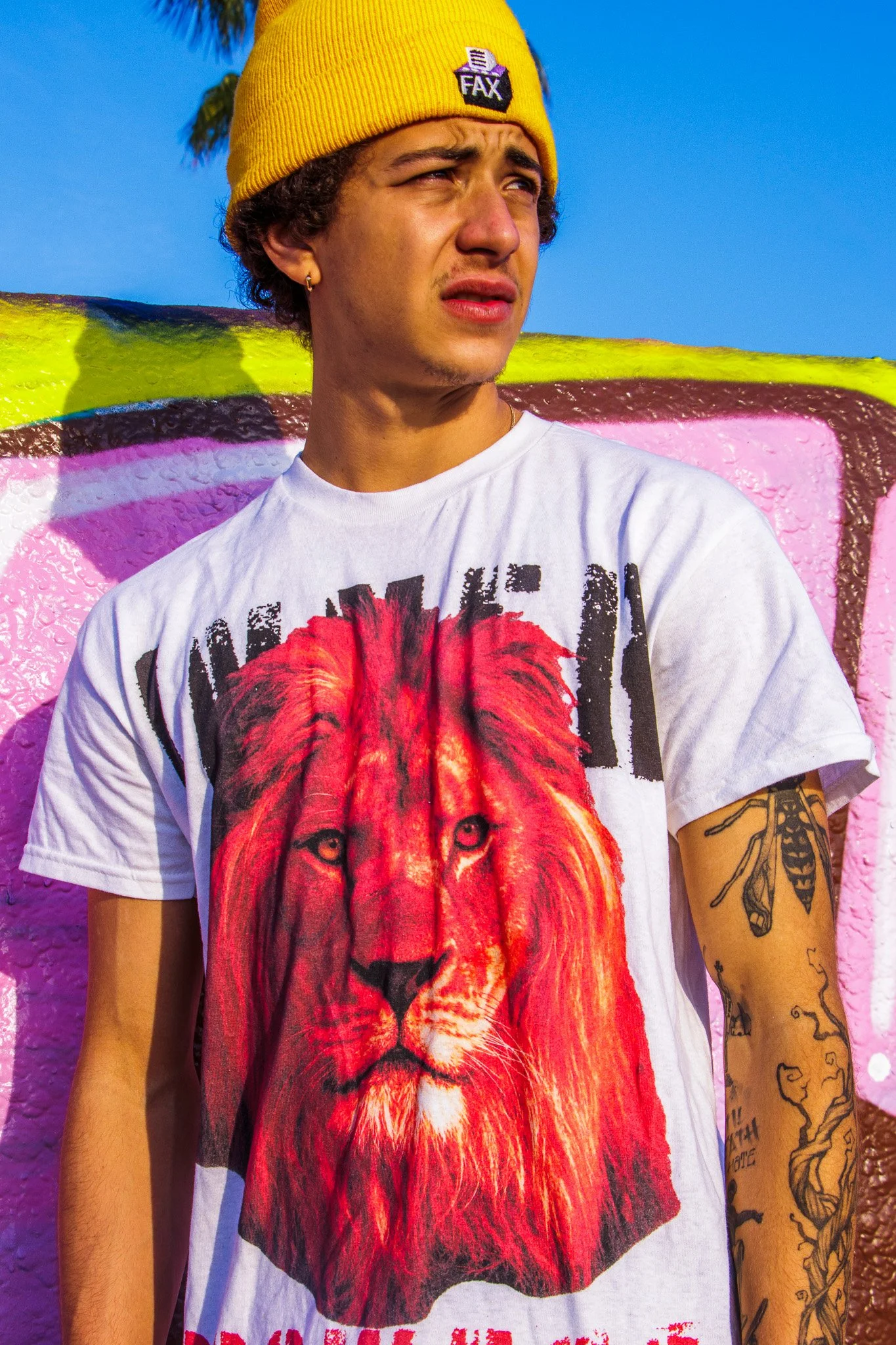 Young man with curly hair wearing a yellow beanie with a small logo, a white t-shirt with a large graphic of a lion, and tattoos on his right arm. He stands in front of a colorful graffiti wall with pink, yellow, and other vibrant colors, against a c