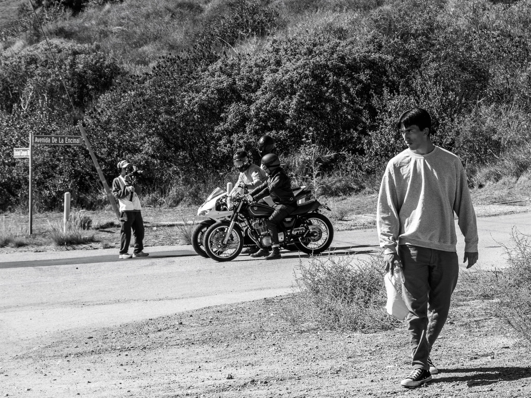 People standing and sitting on motorcycles on a roadside near a bushy hillside, with street signs reading "Avenida De La Encinal" and "Bachillerato Encinal" visible.