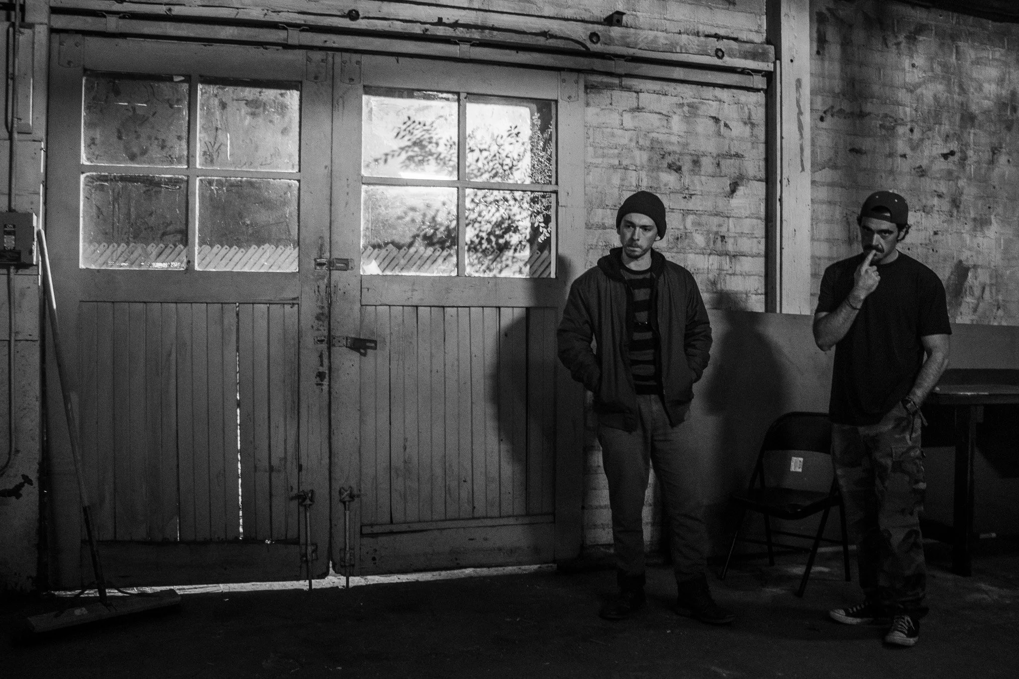 Two men standing inside a garage or workshop, with a large wooden door in the background. One man is wearing a beanie and striped shirt, the other has a backward cap and is touching his lip. The room has brick walls, a chair, and some equipment.