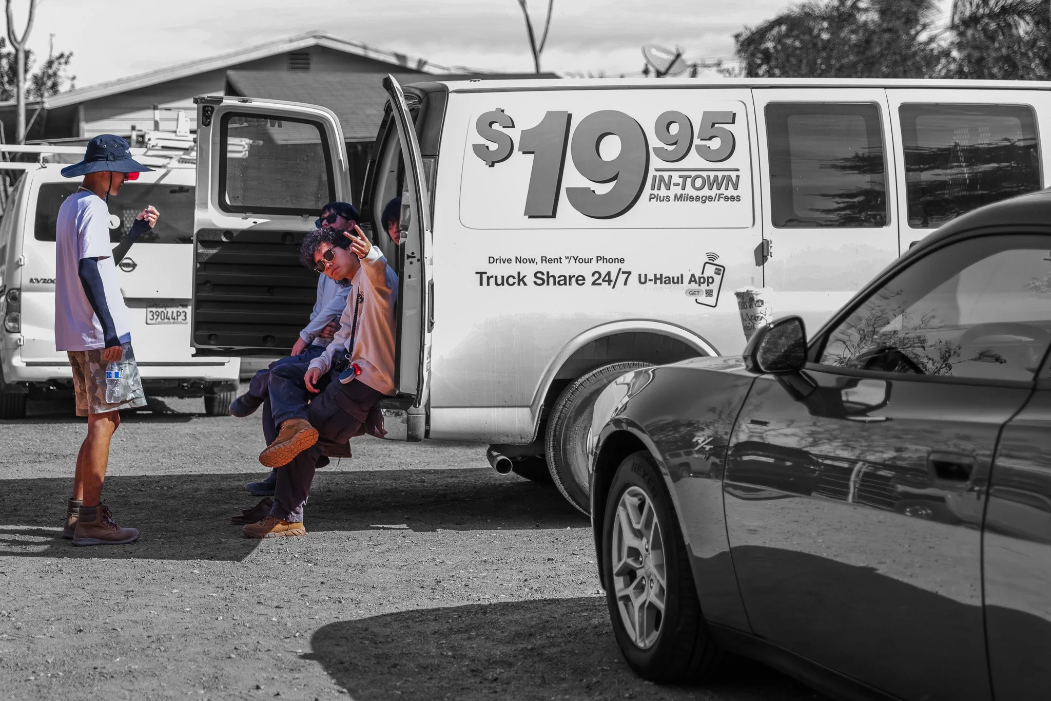 People sitting and standing near a white van with advertisement for truck sharing service, in a parking lot with cars and a building in the background.