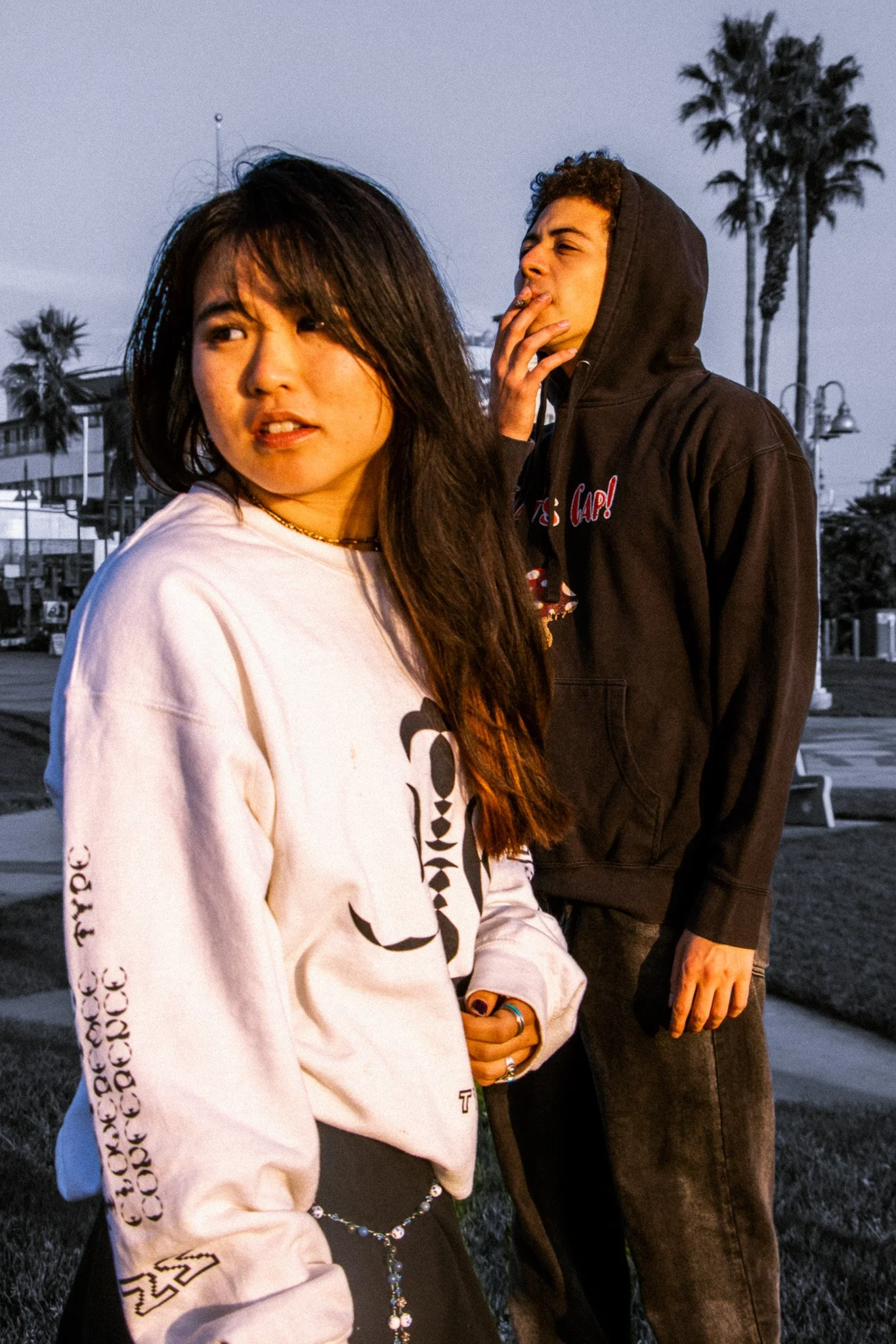 Two young adults, a woman and a man, standing outdoors at sunset or dusk, with palm trees and a cloudy sky in the background. The woman has long dark hair and is wearing a white sweatshirt with black text, and the man has curly hair, is wearing a bla