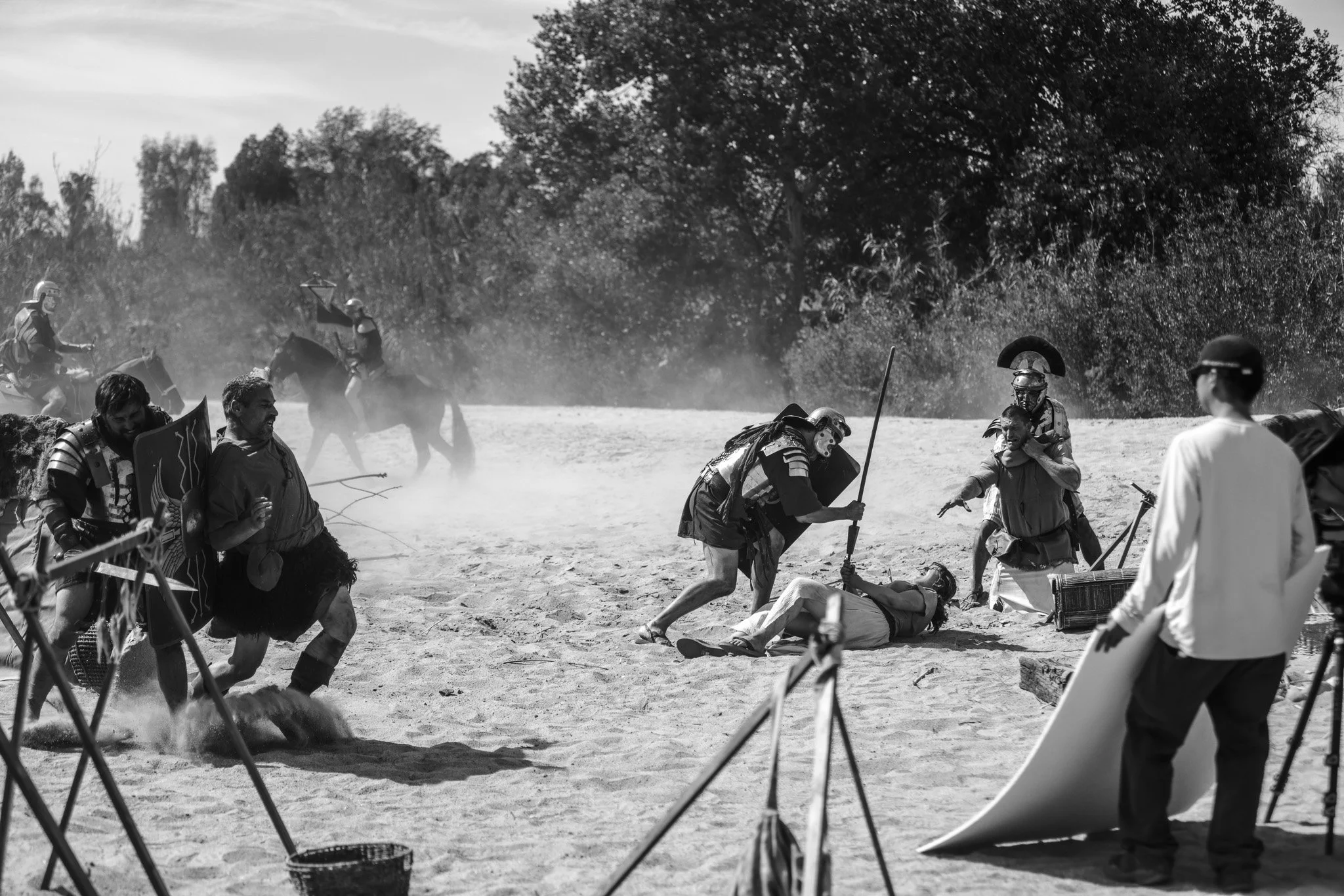 Scene depicting a Roman gladiator attack with fighters, a fallen combatant, and spectators, set outdoors with trees in the background, in black and white.