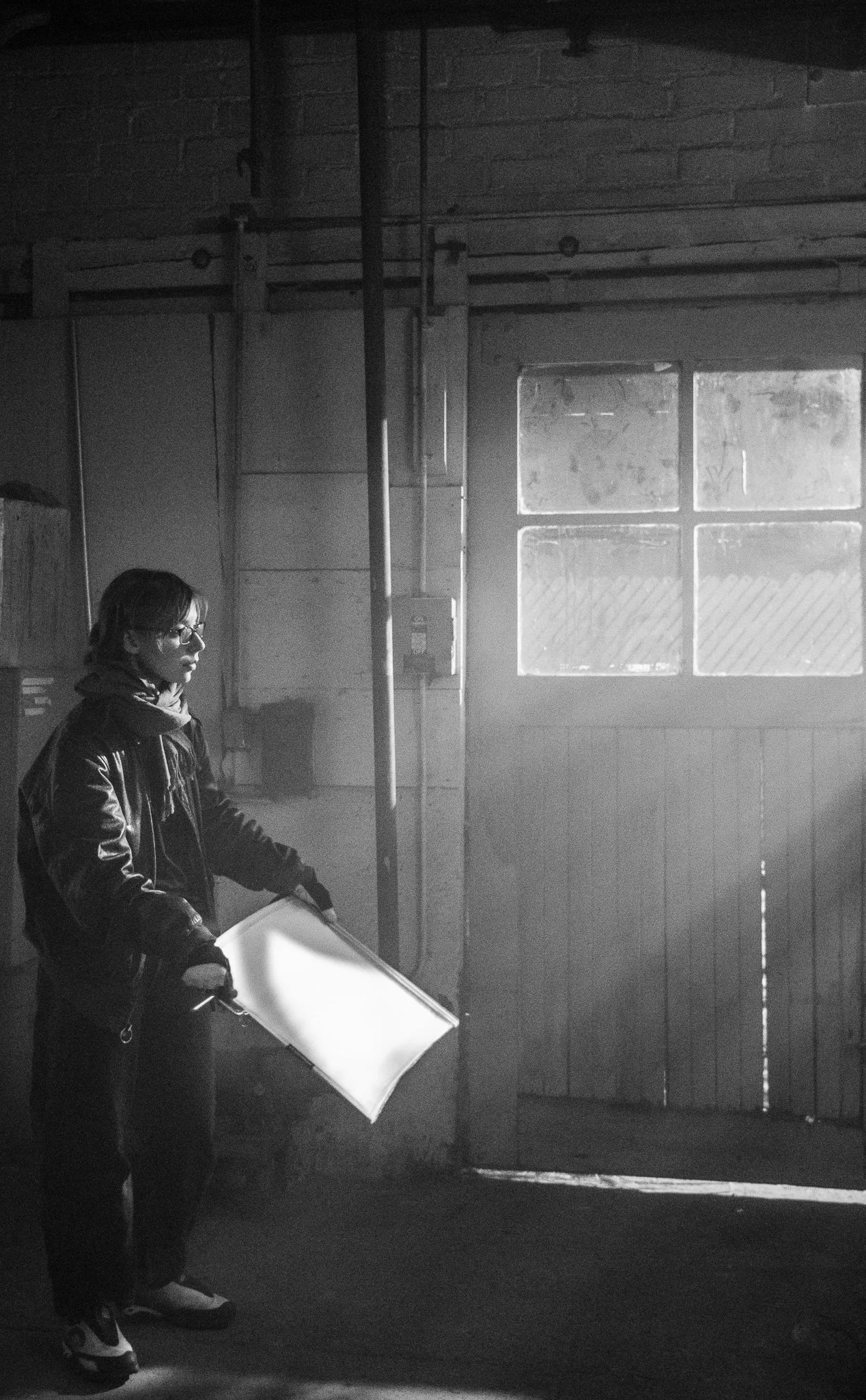 A person wearing glasses and a leather jacket holding a large white object, standing in a dimly lit industrial space with a window and weathered wall.