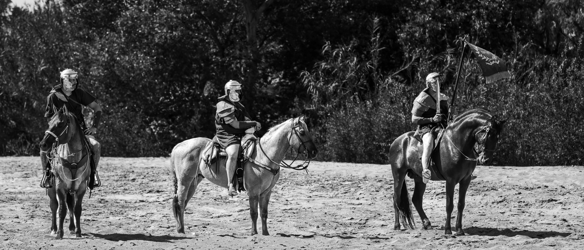 Three horseback riders in medieval-style armor on a dirt field, with trees in the background. One rider holds a flag with a design.