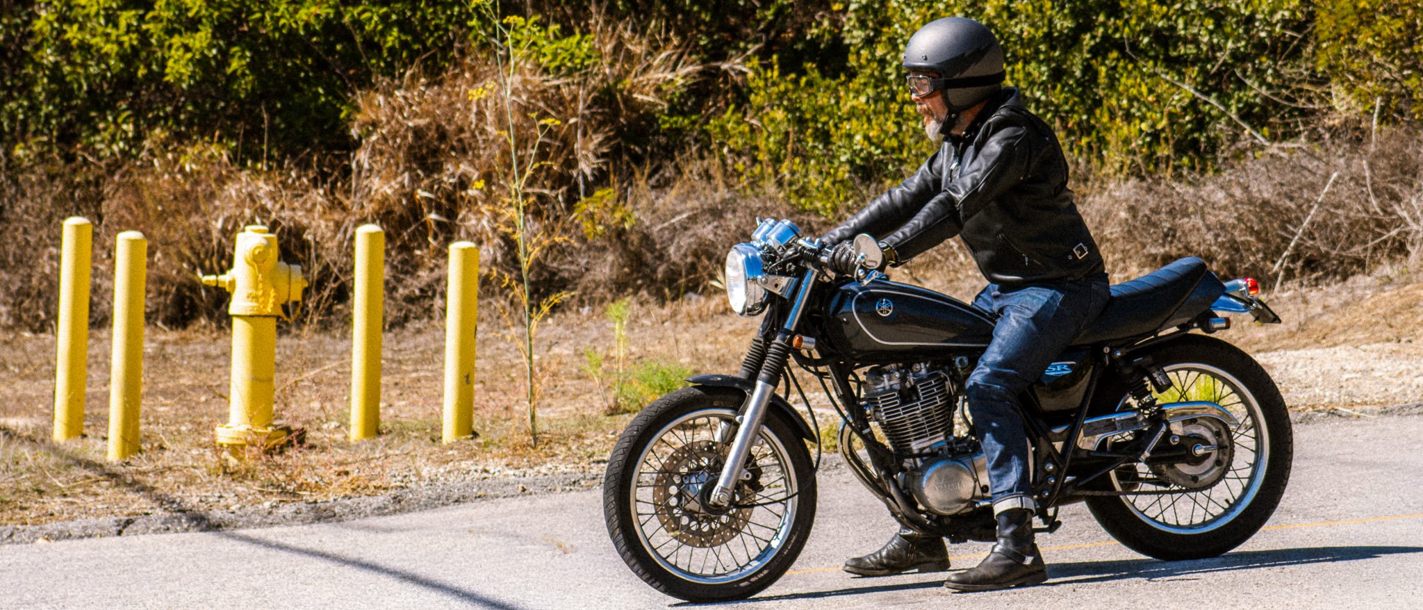 A man wearing a helmet and black leather jacket riding a black motorcycle on a road beside yellow bollards and bushes.