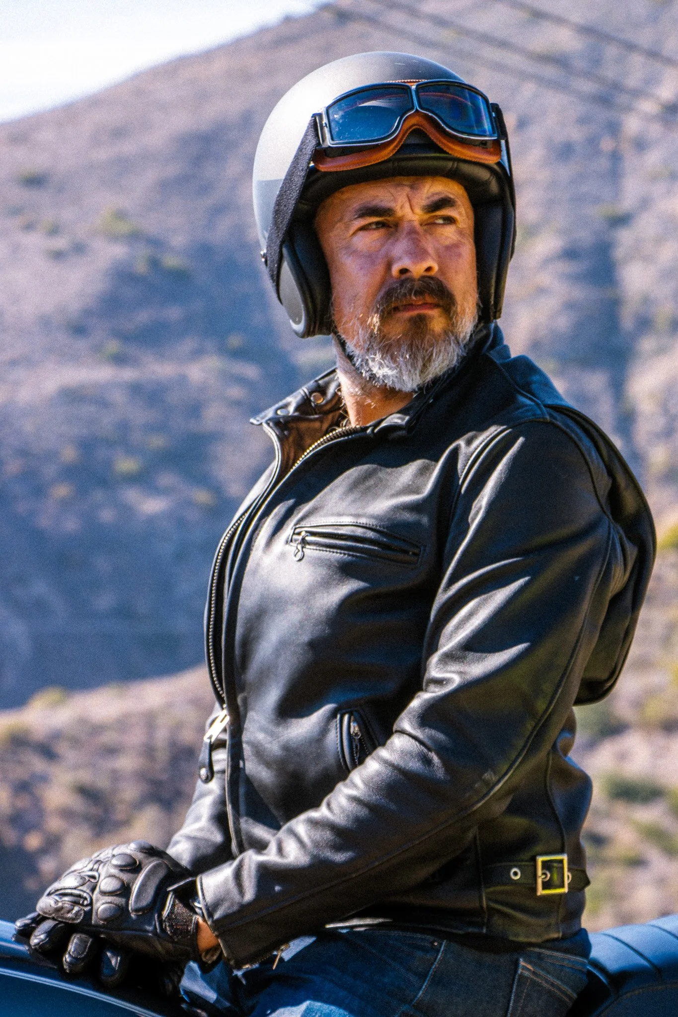 A middle-aged man with a beard wearing a gray open-face motorcycle helmet with goggles on top, black leather jacket, black gloves, and blue jeans, sitting on a motorcycle outdoors with mountains or hills in the background.