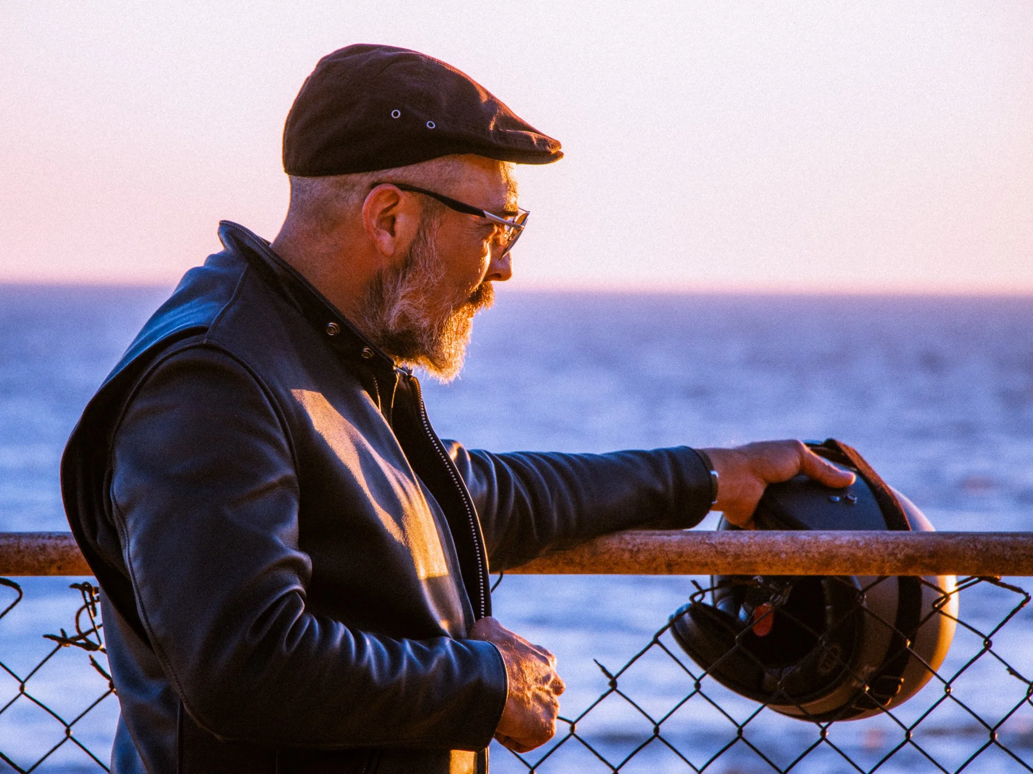 A man with glasses and a beard wearing a black cap and leather jacket, standing outdoors by the water at sunset, leaning on a wooden railing and holding a smartphone near a wire fence.