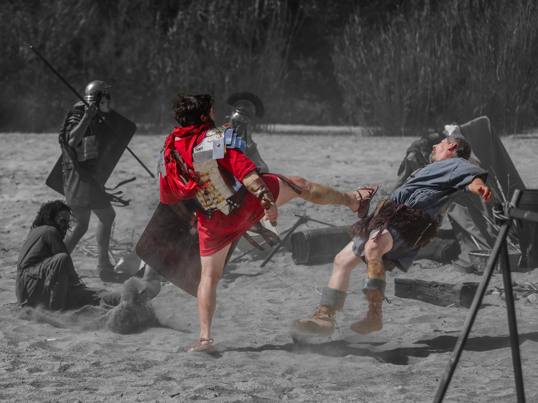 A scene from a movie or reenactment with characters in medieval or fantasy costumes, including a man in a red outfit, a knight with armor, and a person lying on the ground on a sandy or desert-like area. The background is blurred and appears to be ou