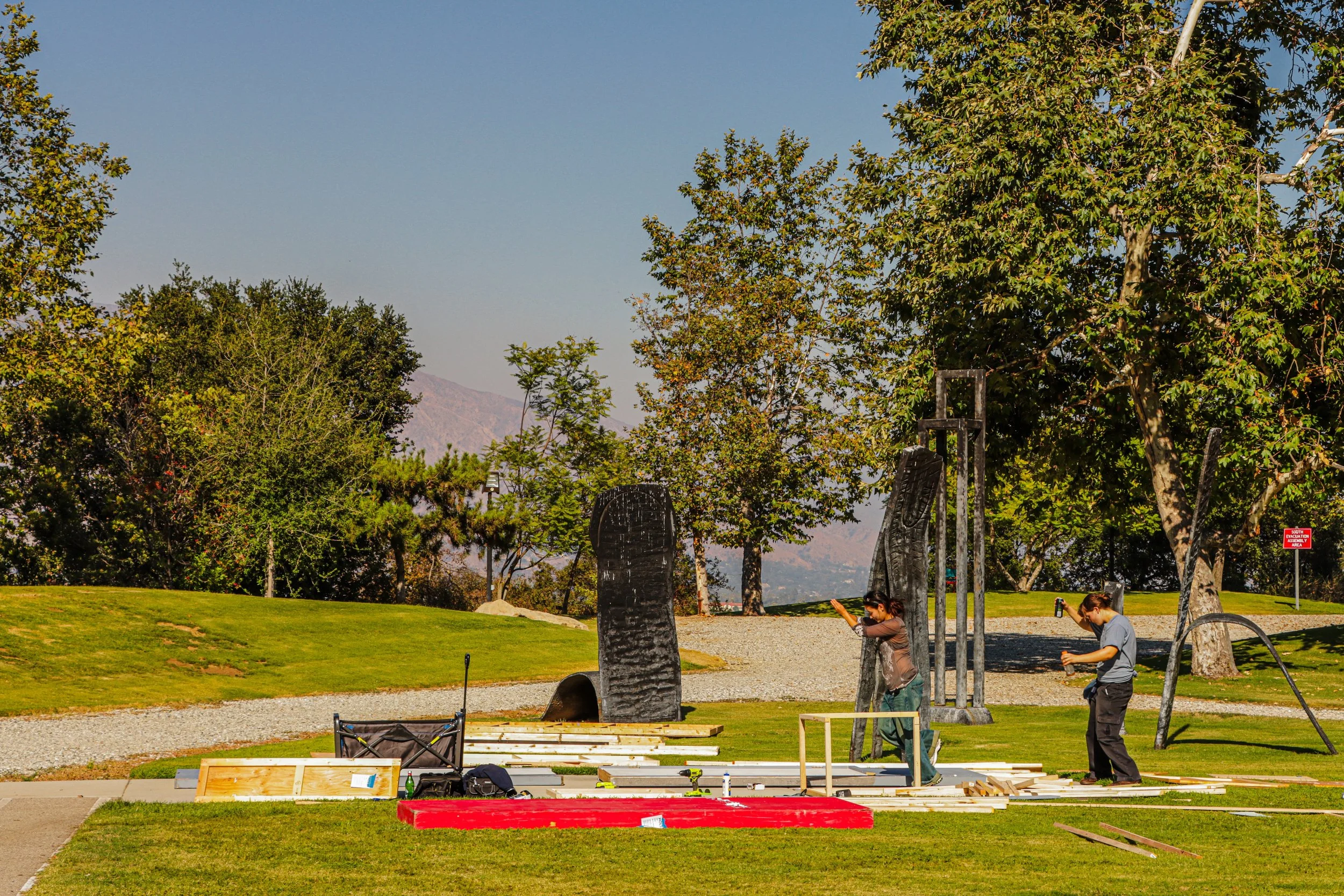 Two people are working on outdoor furniture or structure, surrounded by trees and a grassy area, with construction tools and materials laid out on the ground.