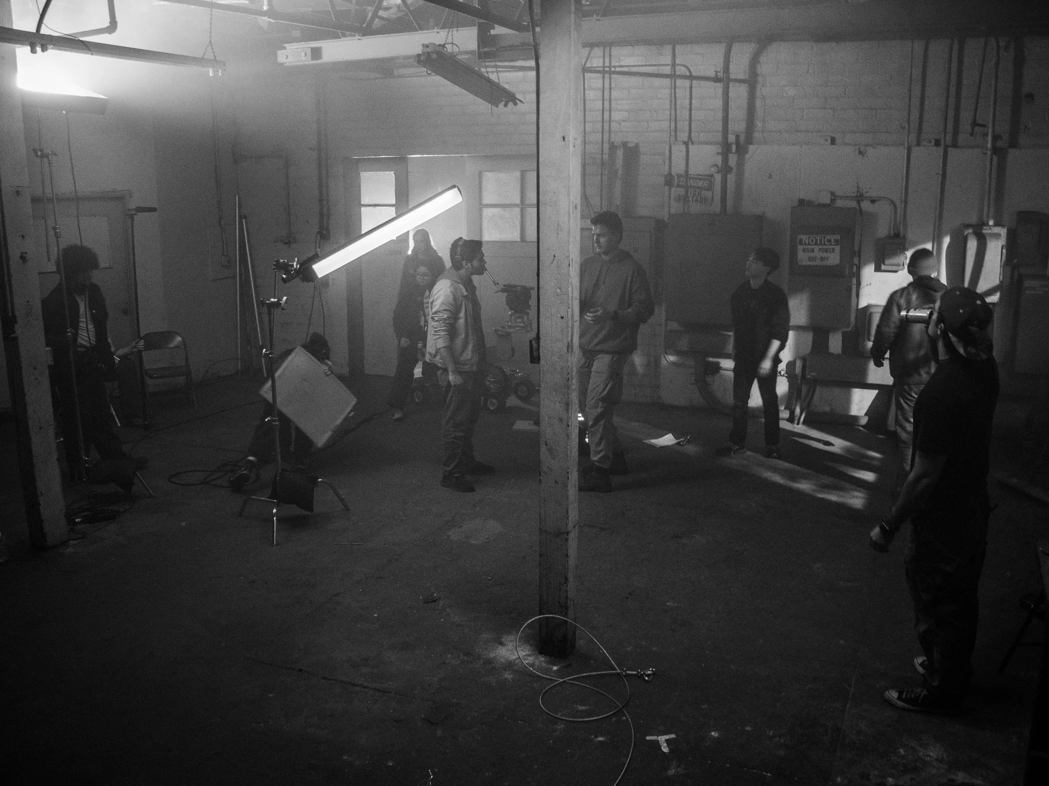 Black-and-white photograph of a film or photography studio with several people working. There is a large light on a stand, some equipment, and people talking or working on their projects in the background.