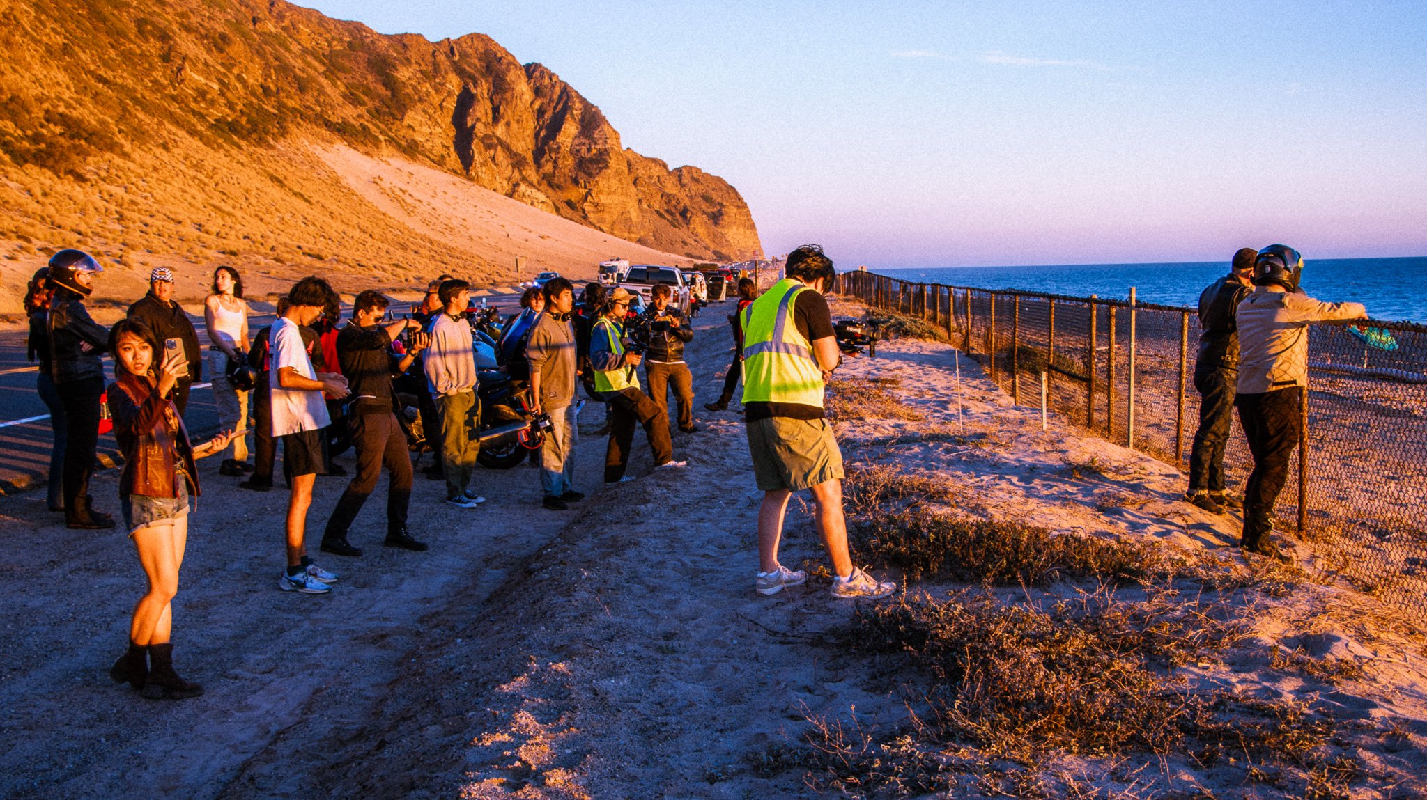 A group of people, some in motorcycle helmets, watching an event along a coastal fence at sunset, with mountains and the ocean in the background.