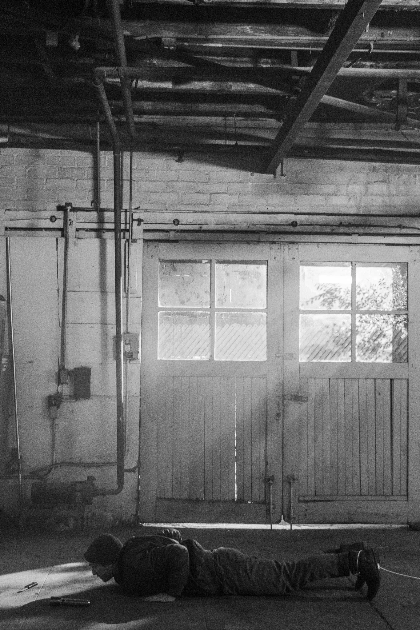 A person lying on their stomach on a mat in a garage or workshop, looking at their phone, with tools and equipment scattered around, and large garage doors with windows in the background.