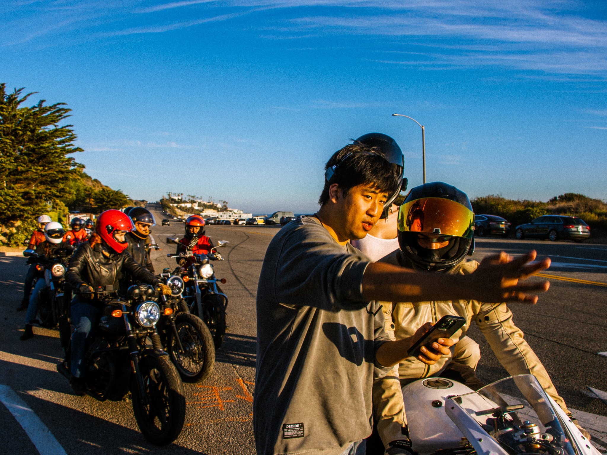 People riding motorcycles in a line on a road near a parking lot, with clear blue sky and some trees in the background, during sunset.