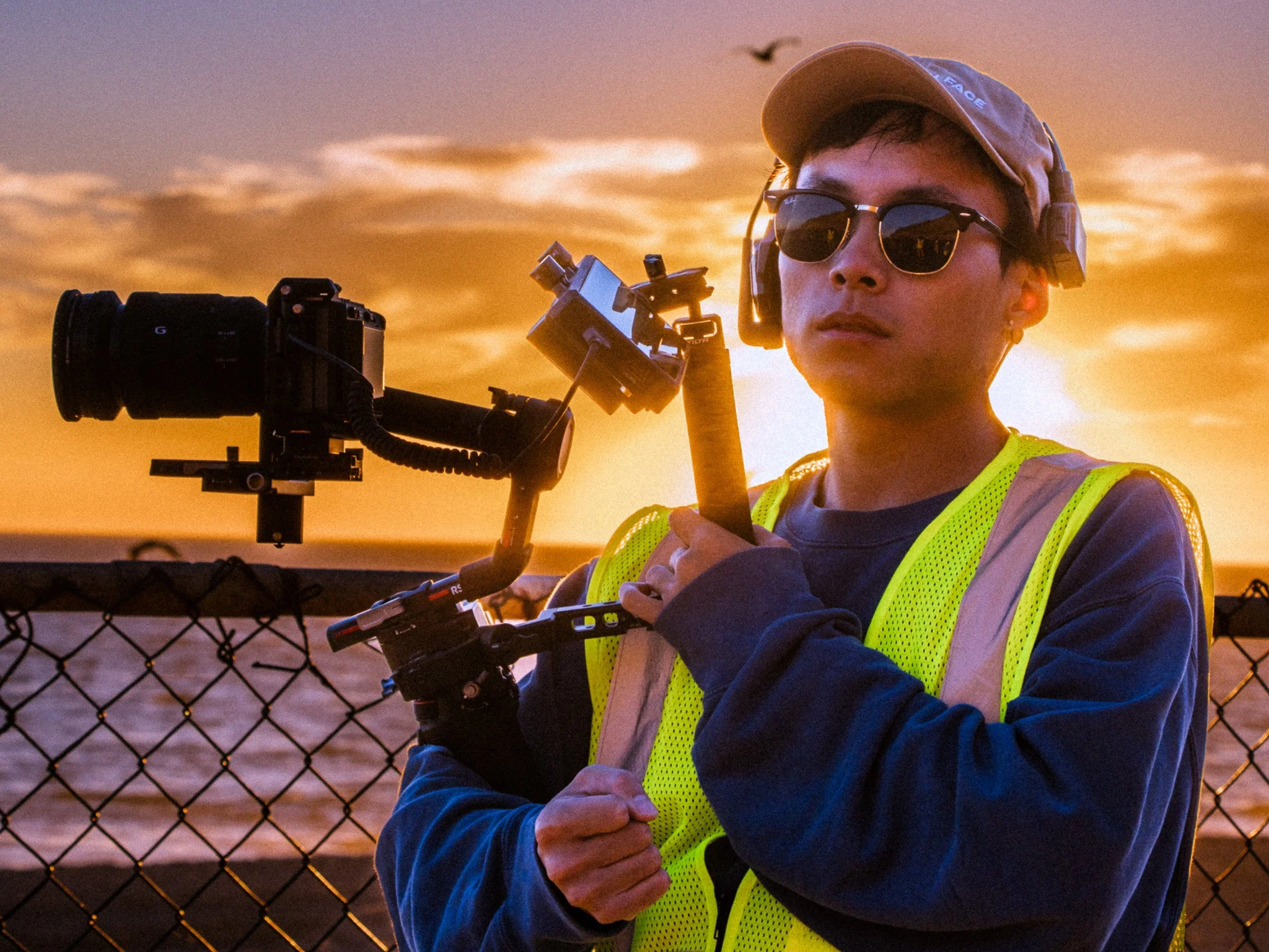 A young person wearing sunglasses, a baseball cap, and a neon yellow safety vest looking into the distance during sunset while holding a camera mounted on a tripod with a camera crane, beside a chain-link fence.