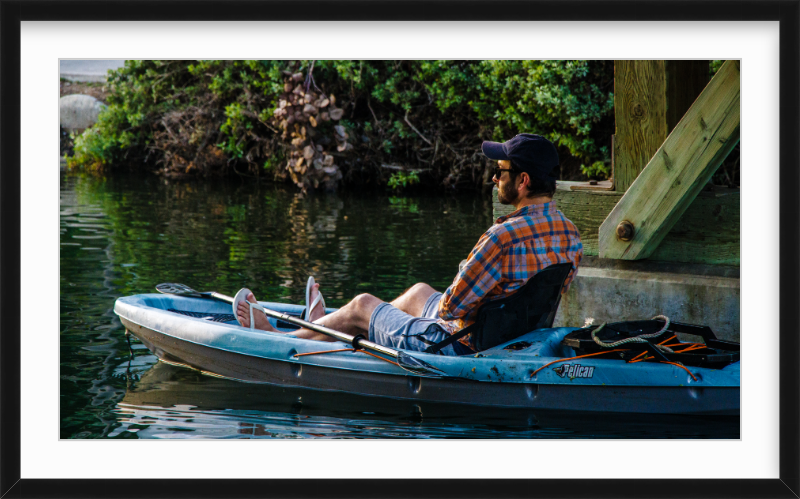 A man in sunglasses and a plaid shirt relaxing on a kayak next to a wooden dock by a calm body of water, with green foliage in the background.