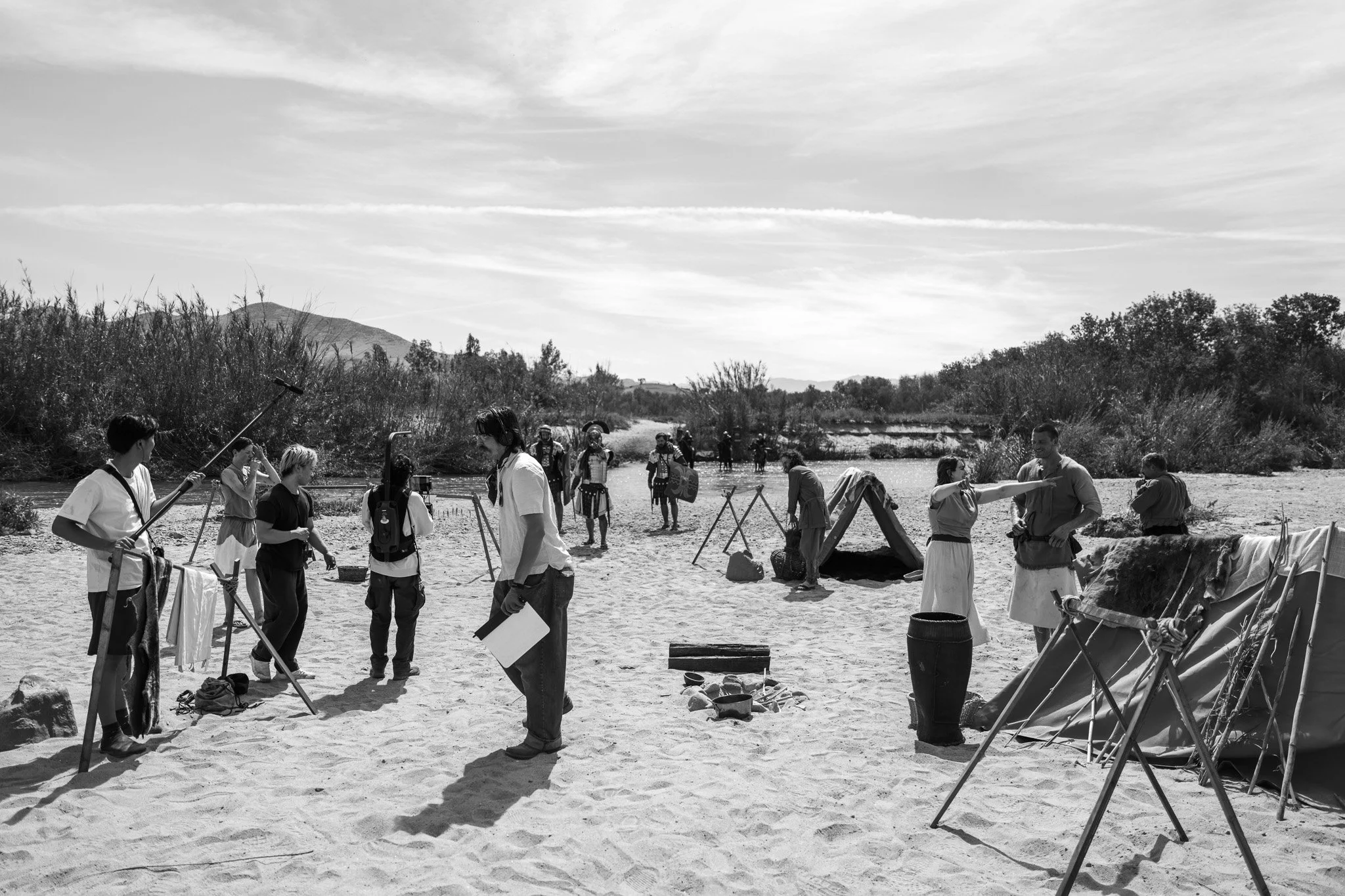 A group of people participating in a historical reenactment or outdoor event on a sandy riverbank, with some individuals dressed in historical clothing and others in modern attire, surrounded by trees and open landscape.