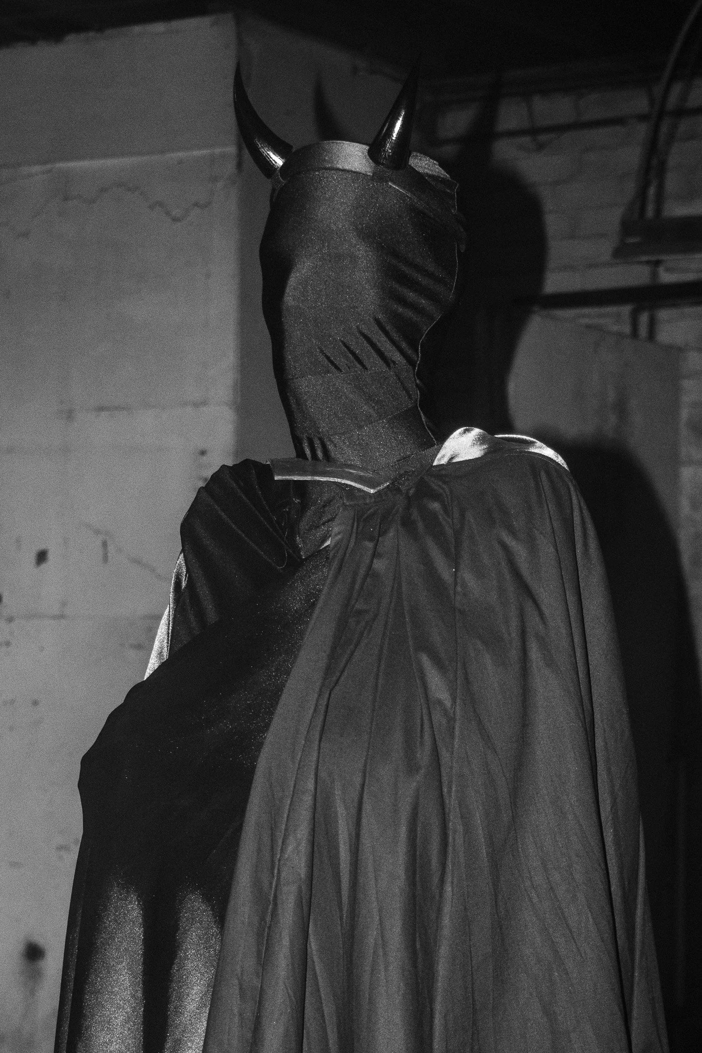 A person wearing a black hoodie with a large, dark cape with vertical lines, and a black mask with two prominent horns, standing in a dimly lit room with brick and concrete walls.