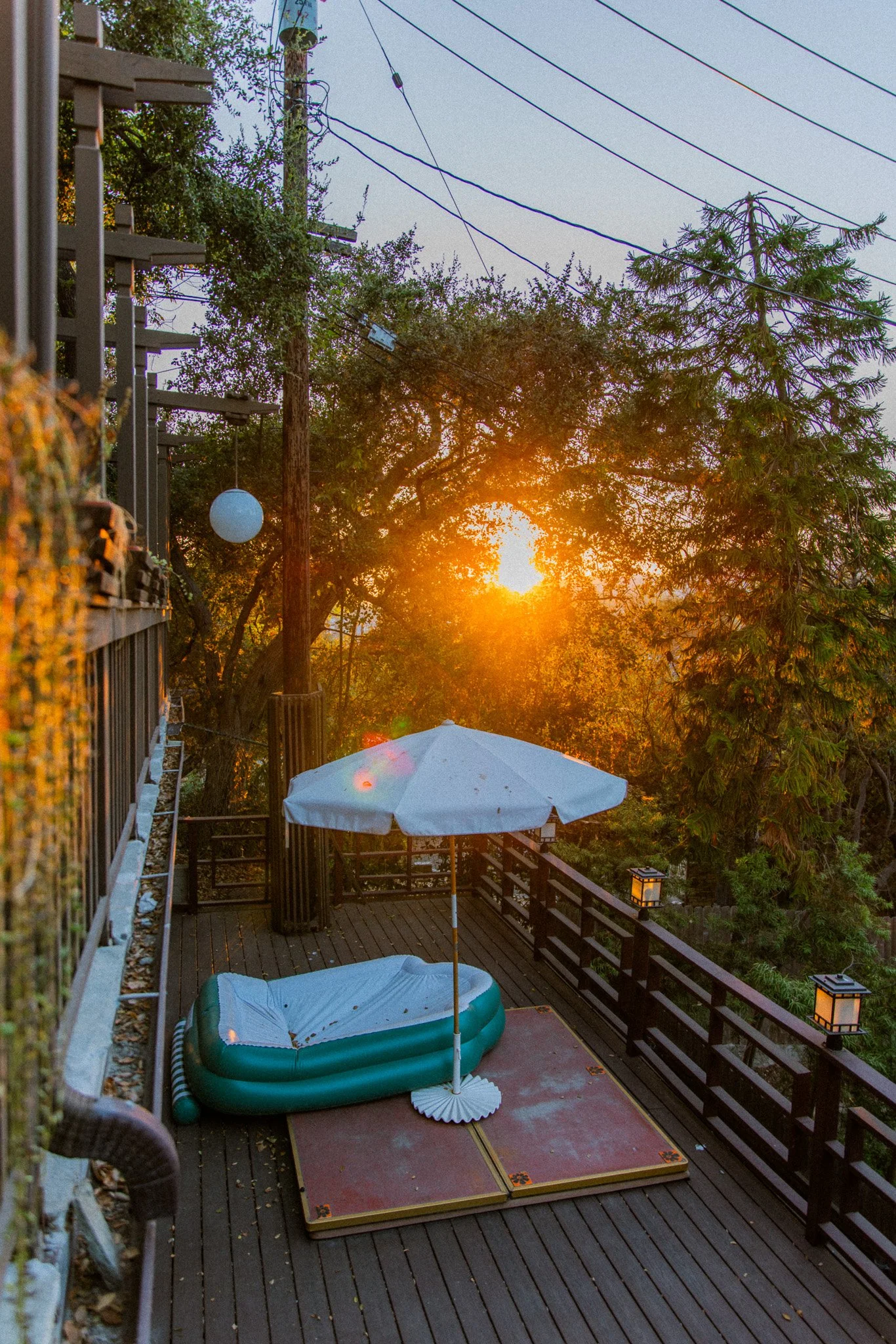 A backyard deck during sunset with a hot tub, patio umbrella, and a striped outdoor rug. There are trees, string lights, and a wooden railing surrounding the deck.