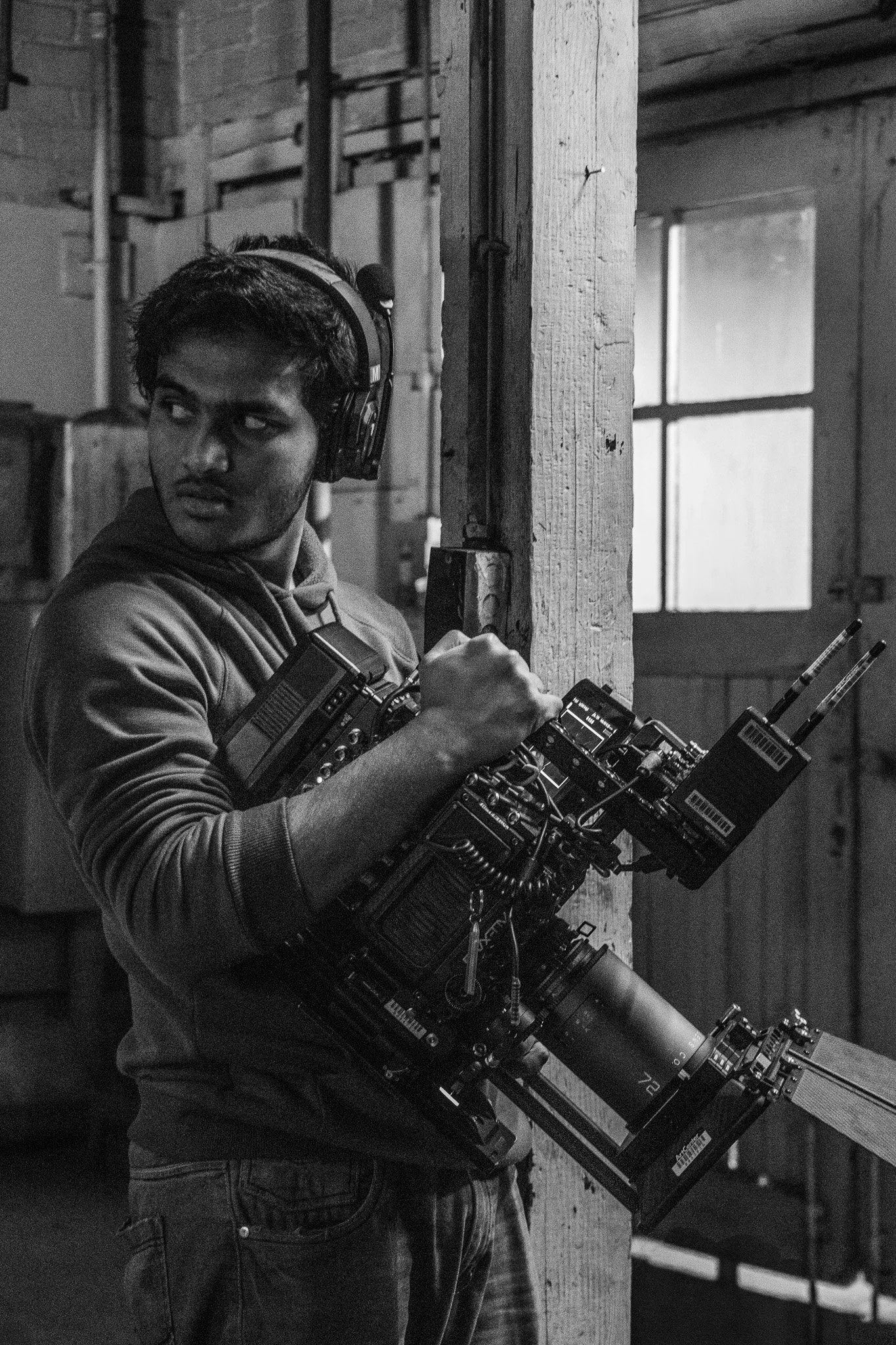 A person wearing a hoodie and jeans, holding a large film or camera equipment, standing indoors against a wooden beam in a rustic setting, with a window and other people in the background.