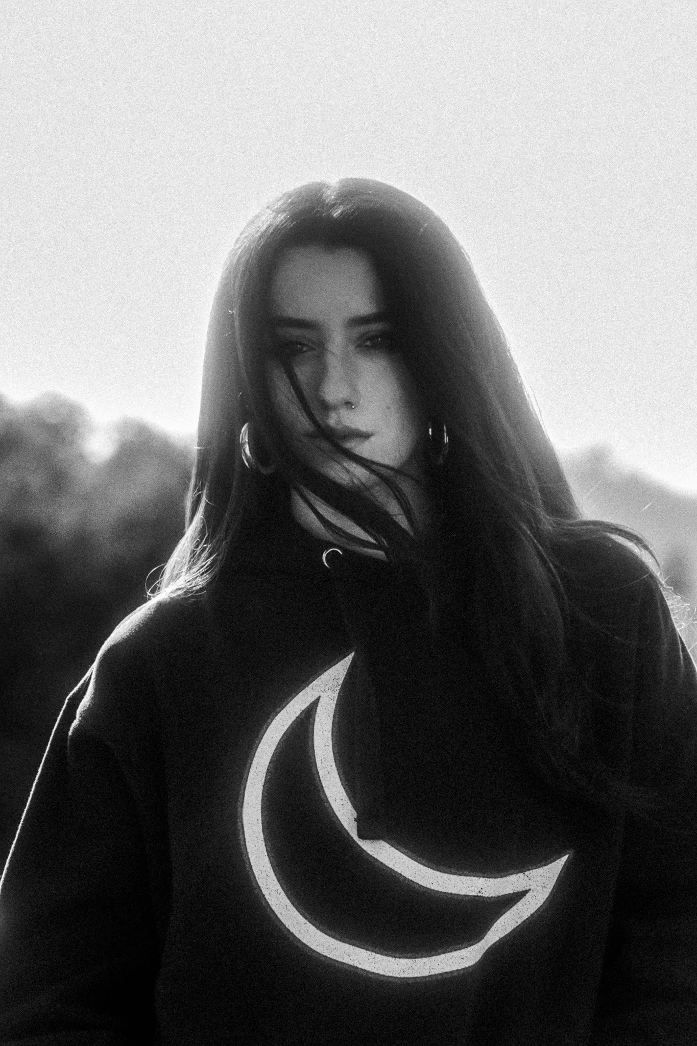 A black-and-white photo of a woman with long dark hair, wearing hoop earrings and a piercing on her nose, outdoors with a blurred background of trees and sky. She is wearing a shirt with a circular moon design.