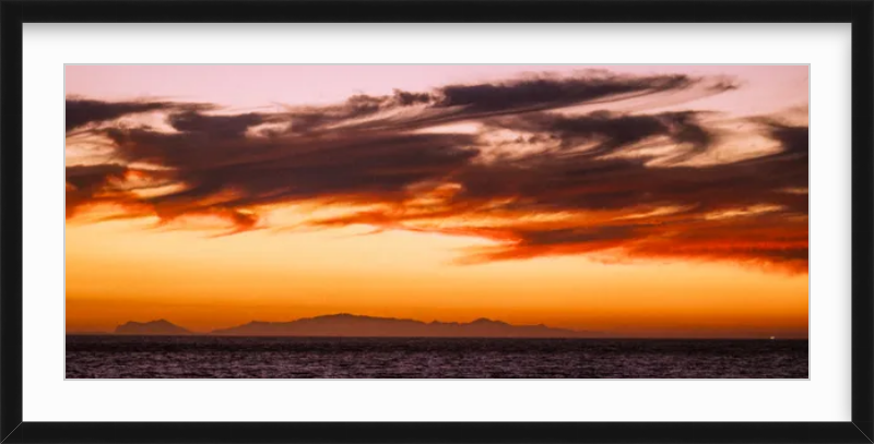 Framed photo of a colorful sunset over the ocean, with dark clouds and a distant silhouette of mountains.