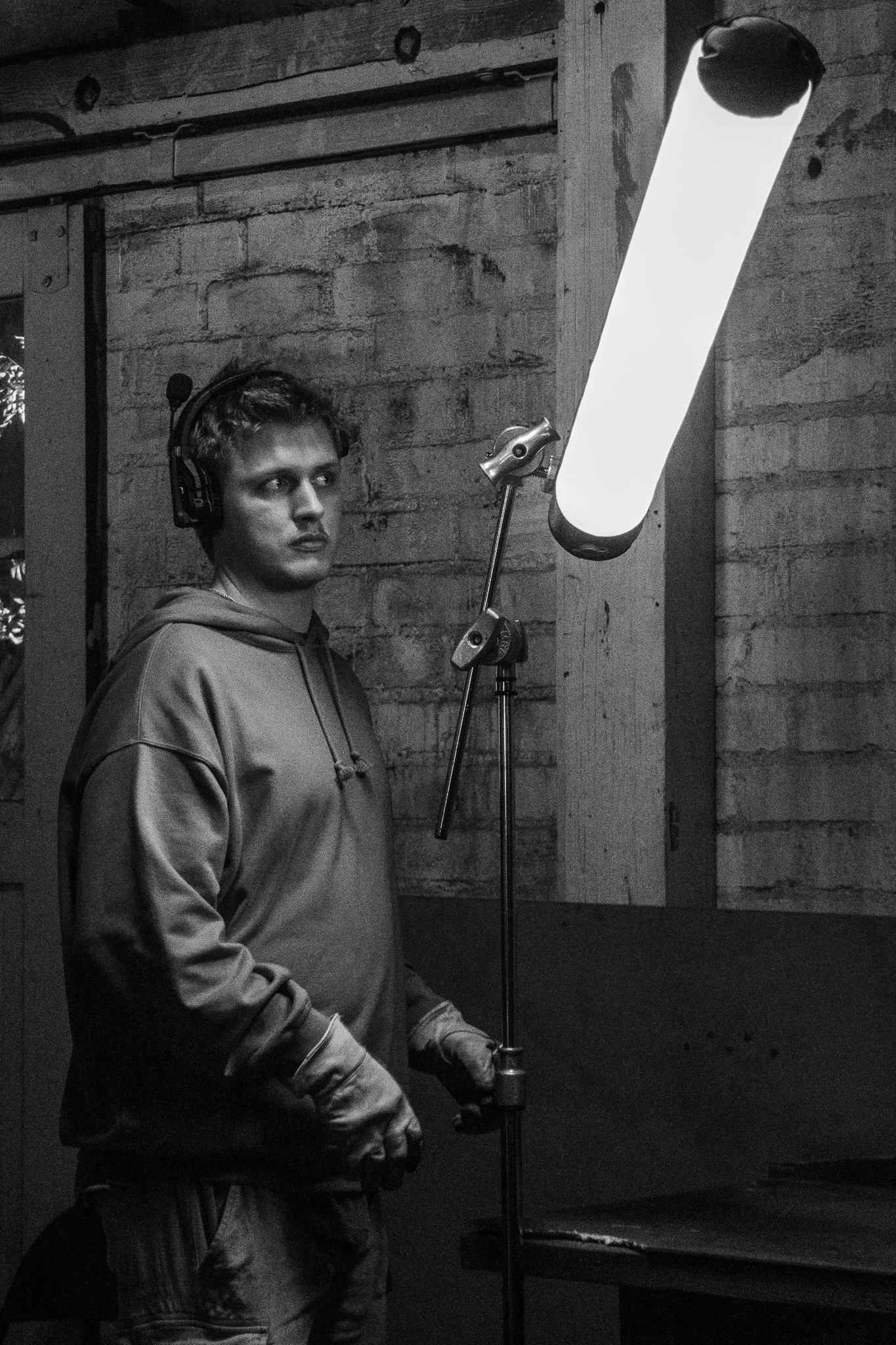 A young man wearing a hoodie and gloves stands in a dimly lit space, wearing a headset with a microphone, next to a tall bright light on a stand, against a rough wooden wall.