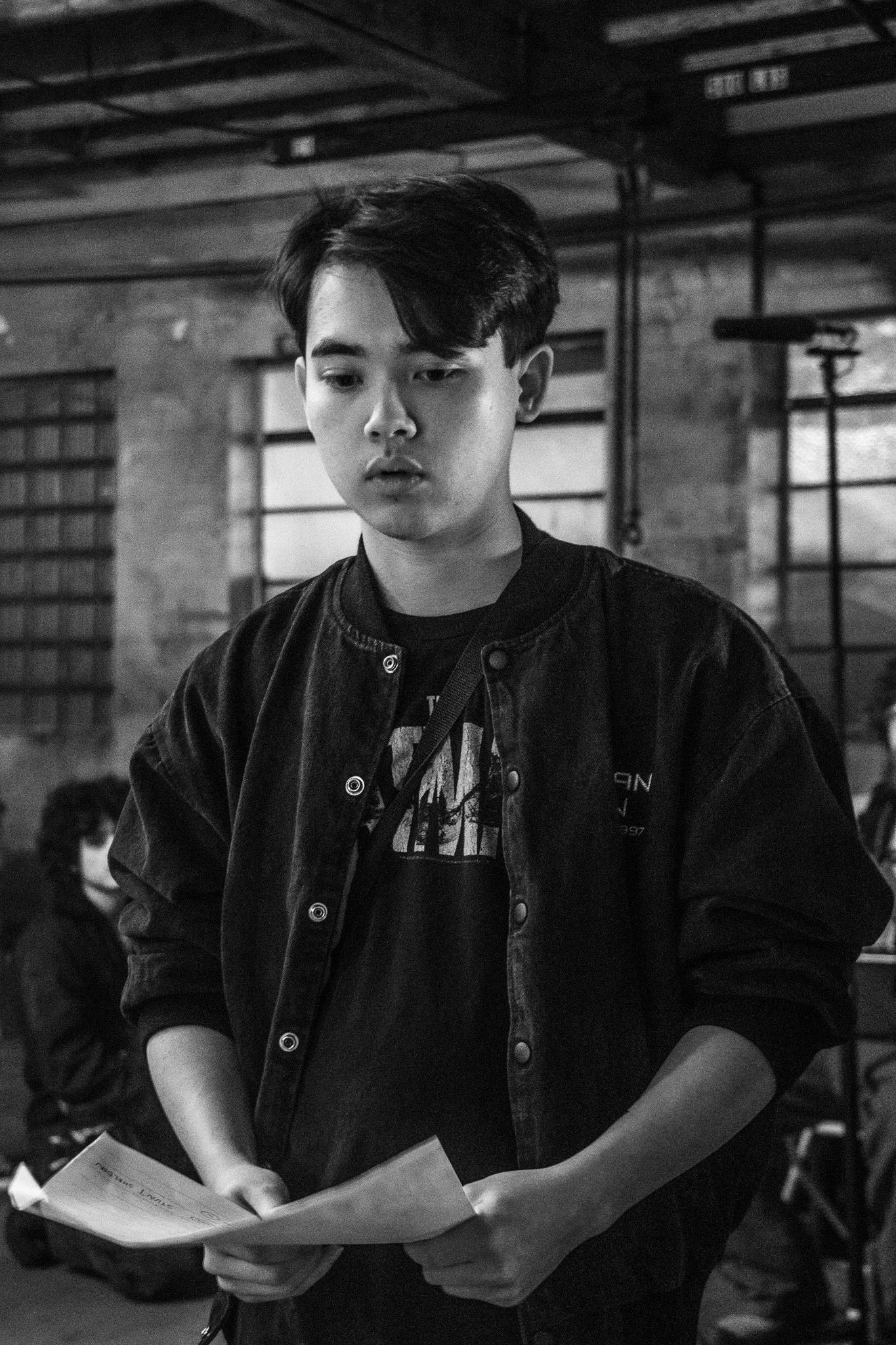 A young man with black hair wearing a black jacket and graphic T-shirt, holding papers, standing in an industrial-style space with brick walls and large windows, black and white photograph.
