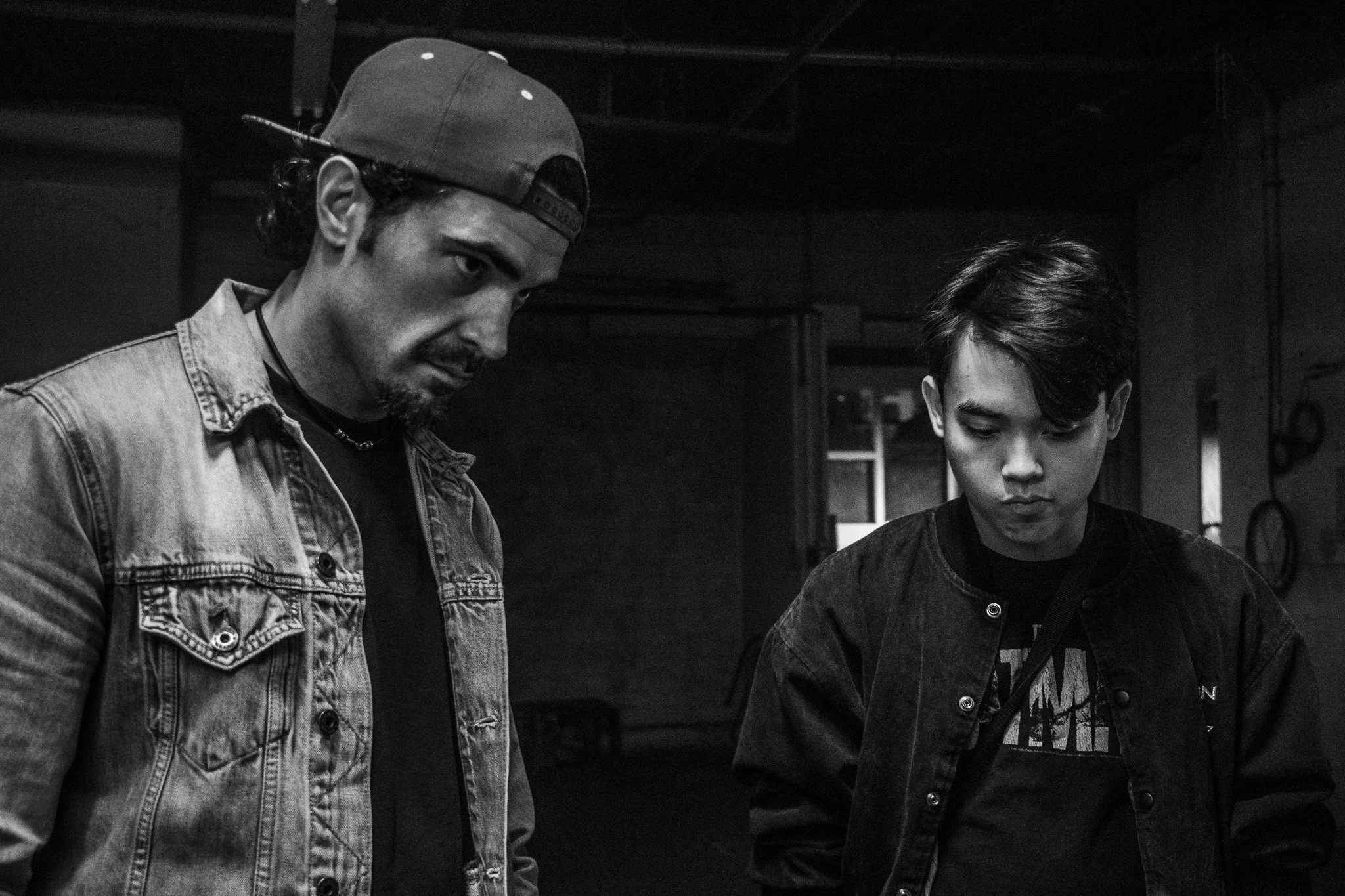 Two young men are looking at something outside the frame in a dimly lit room. The man on the left is wearing a denim jacket and a backward baseball cap. The man on the right has short, styled hair and is wearing a dark jacket with a printed shirt und