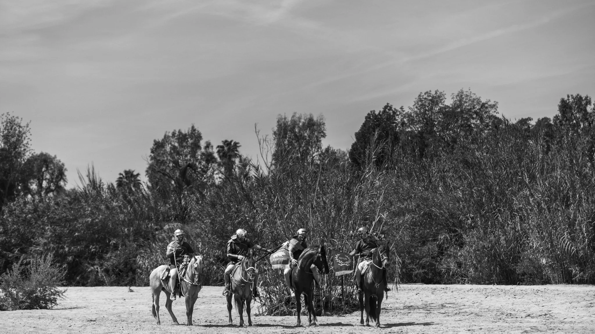 A group of horseback riders crossing a river in a natural outdoor environment with tall trees and bushes in the background, in black and white.